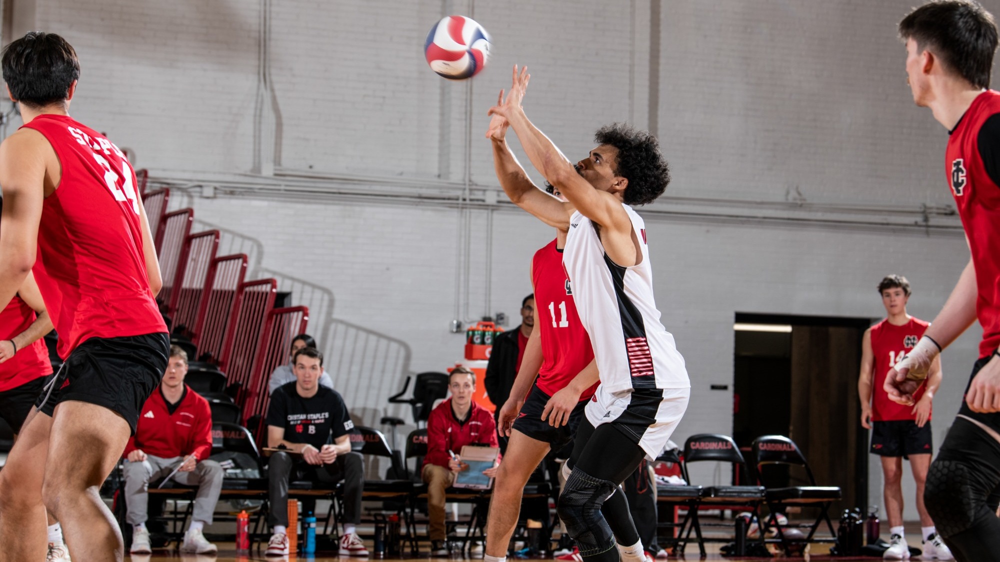 Jonathan Brown sets a ball using two hands for a teammate during a match while wearing a white colored jersey, juxtaposed by the red jerseys around him because he is the libero.