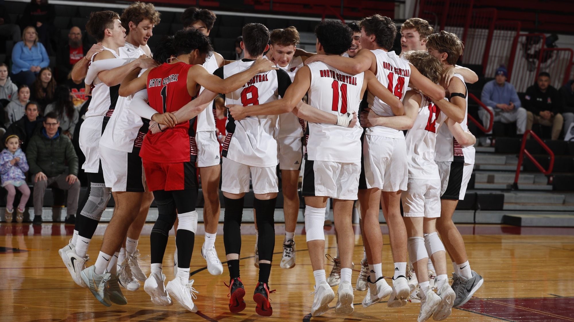 The members of the men's volleyball are in a circle with their arms draped over the person's shoulders to their left and right. While doing this, the team is leaping up from the ground while chanting as a part of a pregame ritual.