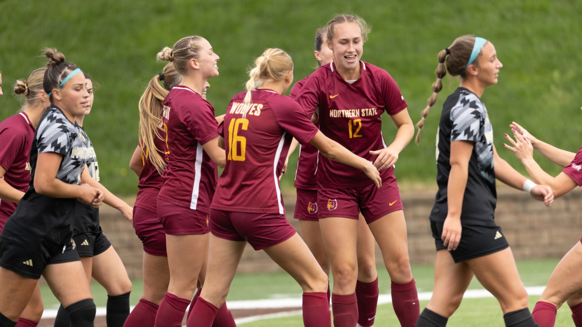Northern State soccer players in maroon uniforms gather together on the field during a match as opposing players in black uniforms walk nearby.