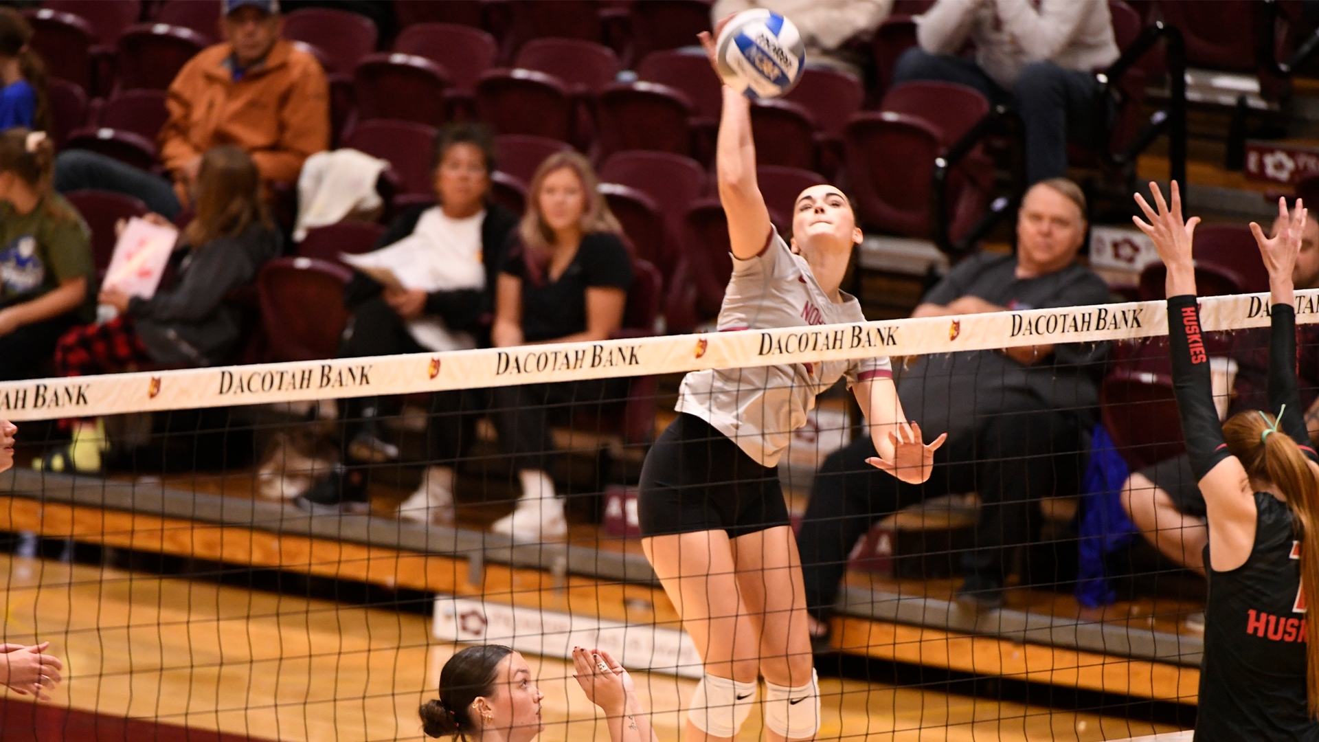 10.23 Volleyball Recap - Lydia Nelson contacts the ball on an attack with defenders in her face as spectators in the background look on