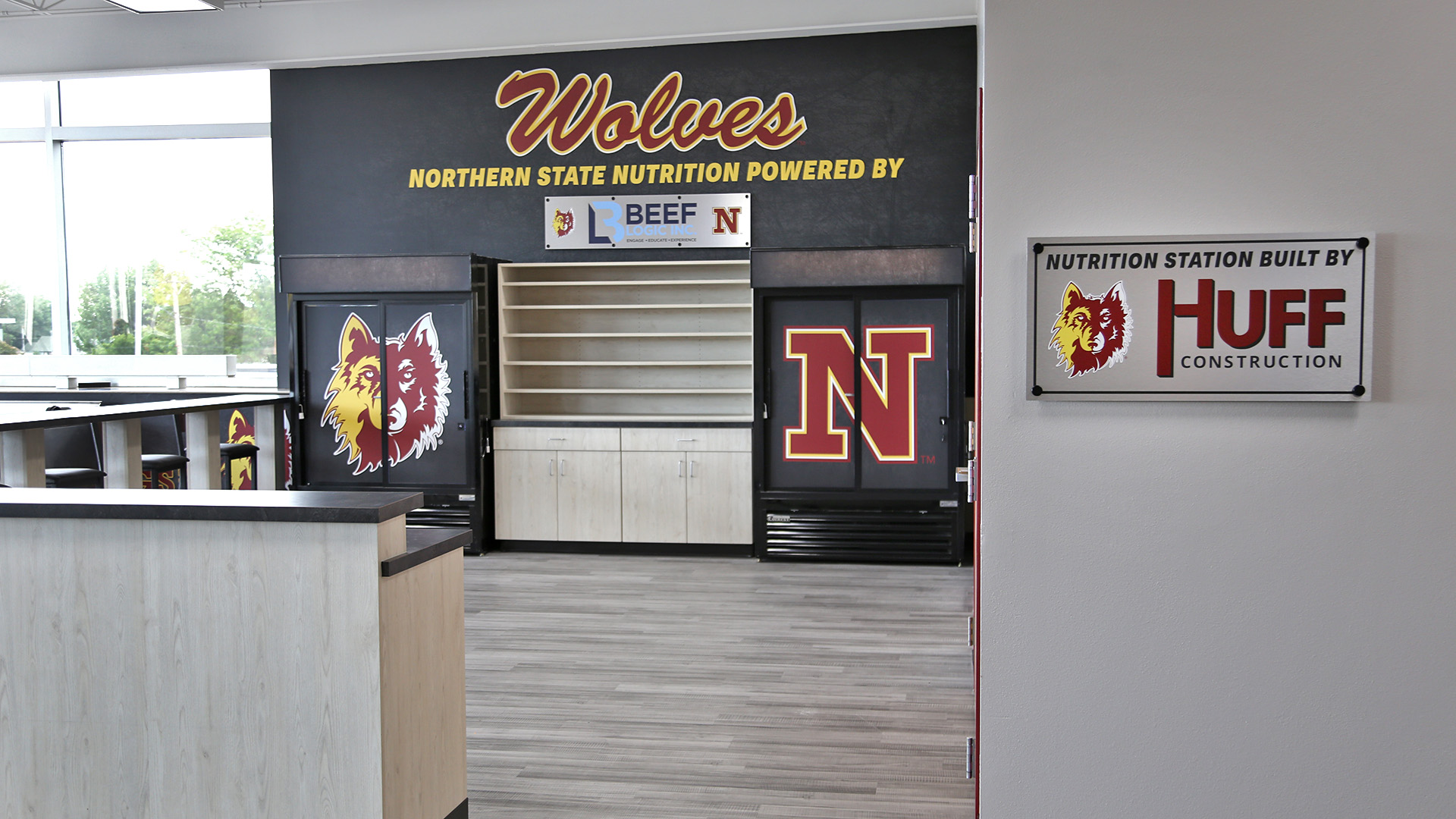The entrance of the NSU nutrition station featuring two vinyl wrapped fridges, countertop seating, and a storage shelf,  as well as wall graphics behind. 