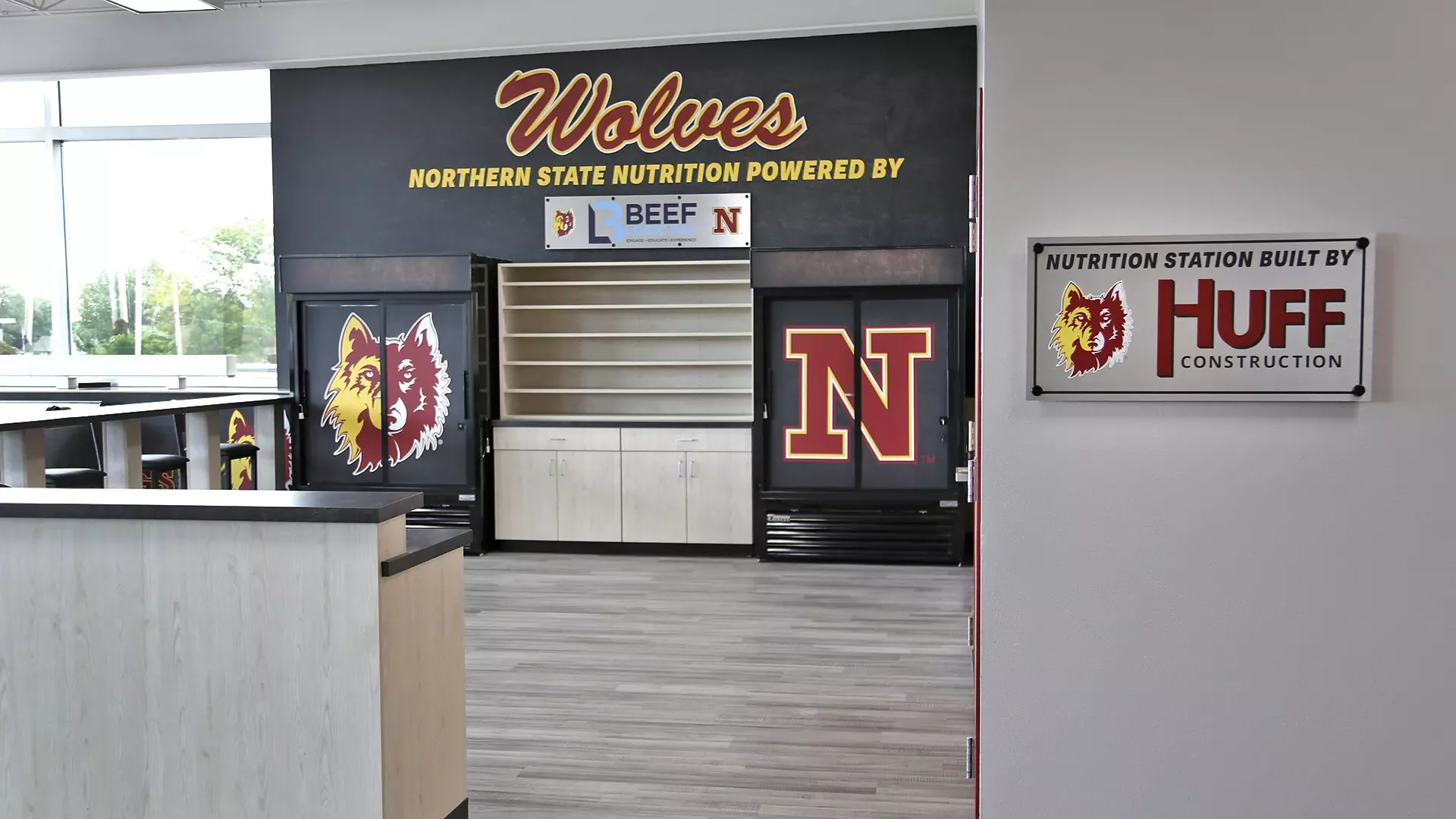 The entrance of the NSU nutrition station featuring two vinyl wrapped fridges, countertop seating, and a storage shelf,  as well as wall graphics behind. 