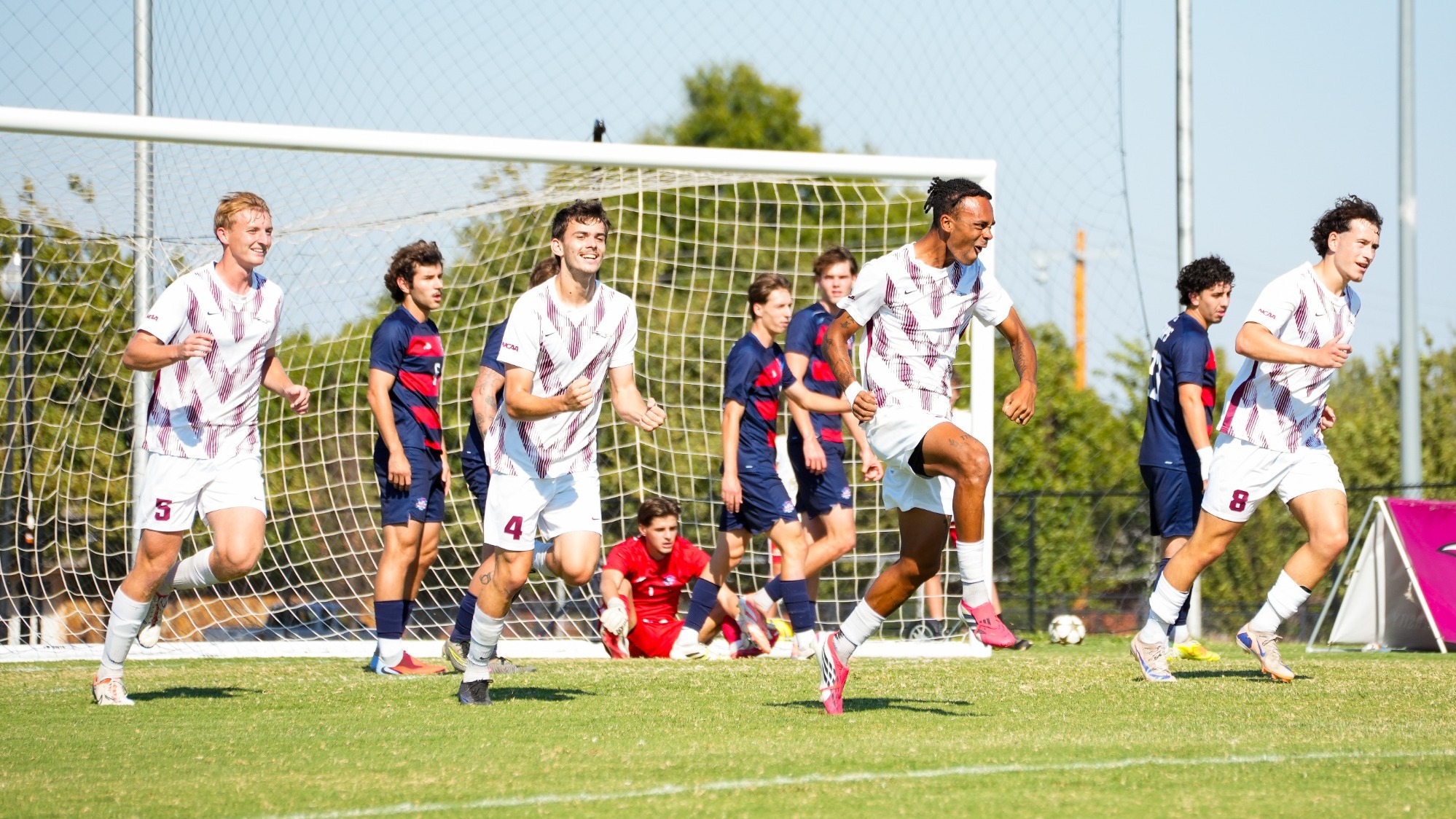 Oklahoma Christian University men's soccer players in white patterned jerseys cheer and run across the pitch in celebration after scoring a goal. Opposing Lubbock Christian players in navy uniforms and the goalkeeper in red watch from near the goal net in the background.