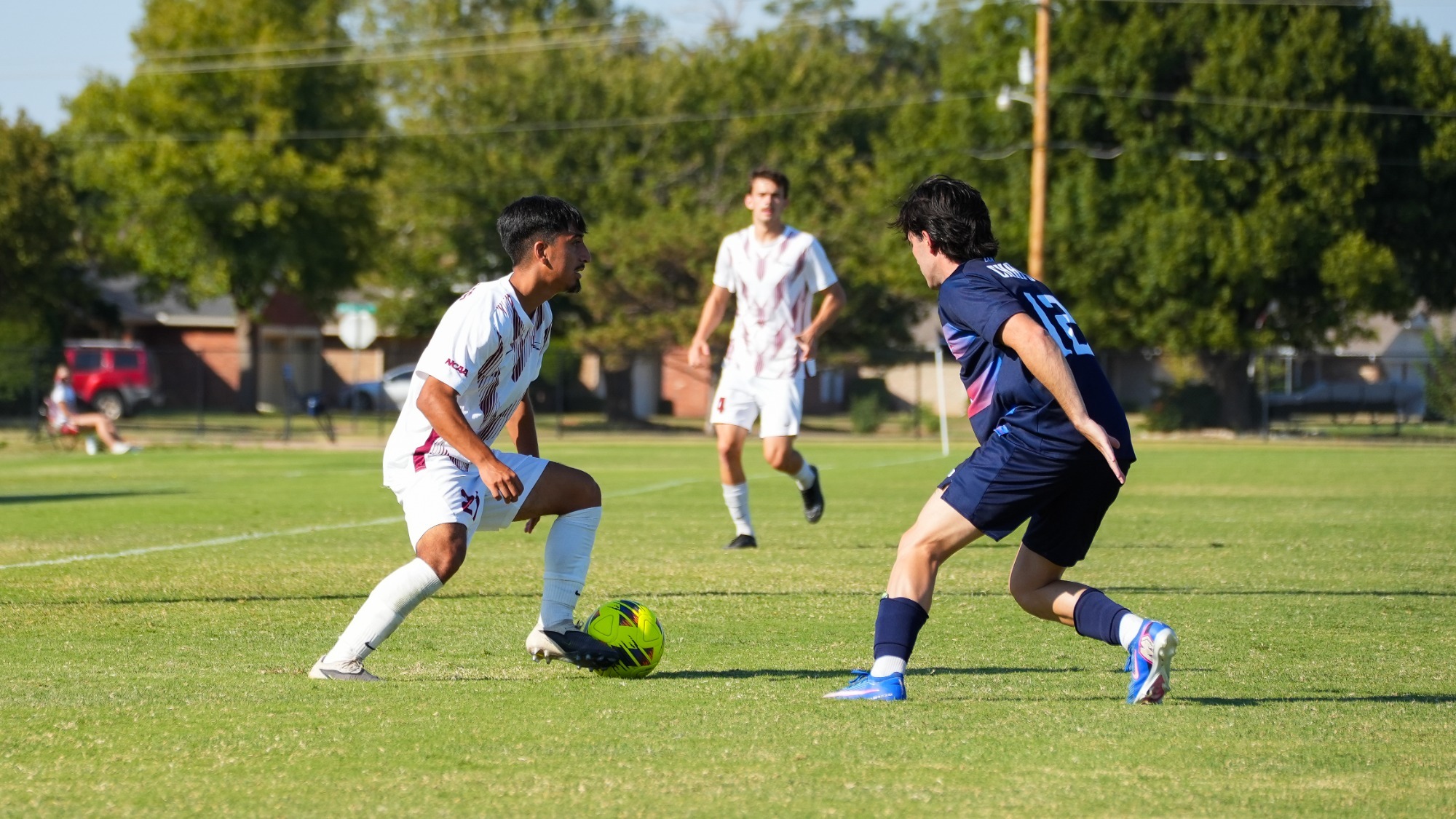 Oklahoma Christian men's soccer player Frederico Palacios (27) in a white and maroon jersey controls a yellow soccer ball on a green grass field while taking on a defender in a dark blue kit.