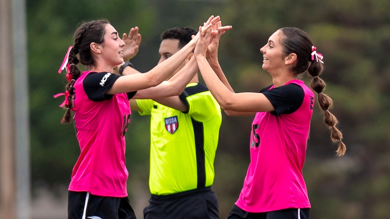 Two Oklahoma Christian women's soccer players in pink jerseys high-five on the field during a match, with a referee standing in the background.