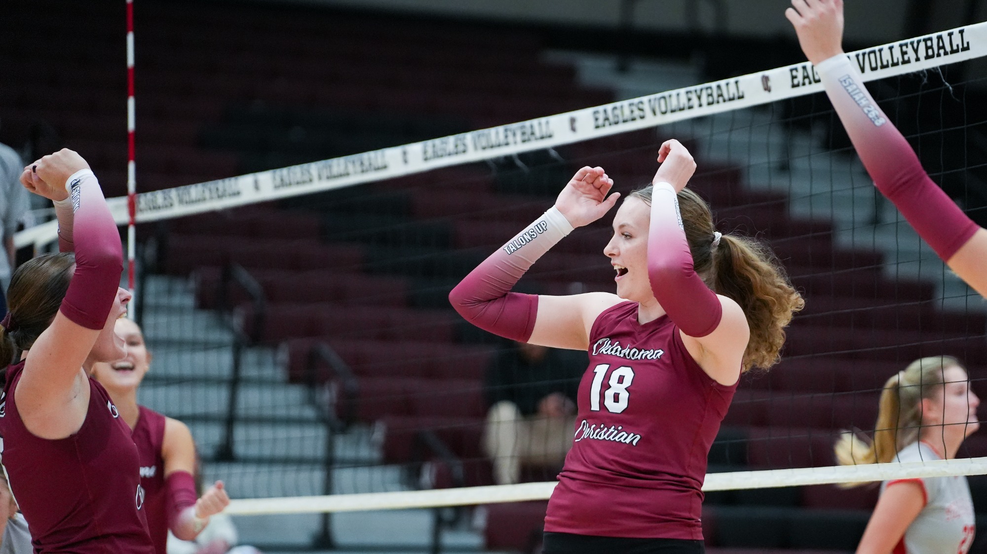 Oklahoma Christian volleyball player Leah Duncan (#18) raising her arms in excitement while celebrating a point with her teammates on court, with the volleyball net in the background.