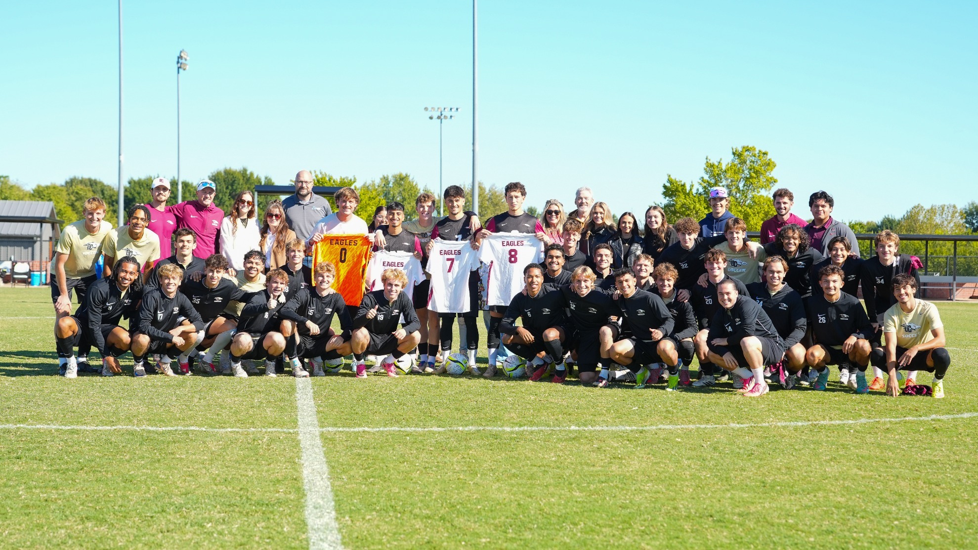 The Oklahoma Christian men's soccer team and coaching staff gather together on the field for a large team photo during a sunny Senior Day celebration. The four senior players stand near the center holding up their framed or displayed team jerseys—including an orange goalkeeper jersey with number 0, and white home jerseys with numbers 7 and 8—surrounded by kneeling and standing teammates on the grass, under a bright blue sky.