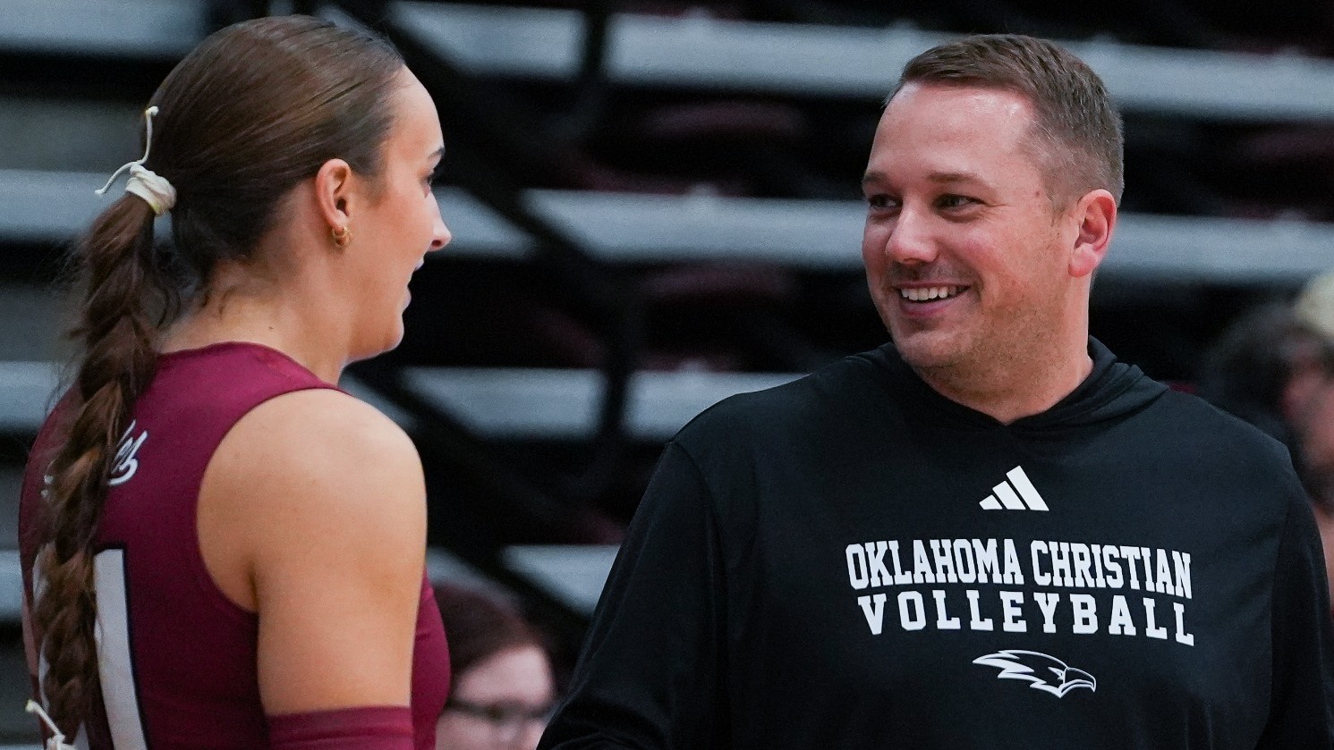 A candid moment showing an Oklahoma Christian Volleyball coach and player interacting and smiling warmly inside a sports arena. The coach, wearing a branded black Adidas hoodie, smiles at a female player in a maroon number 1 jersey with 