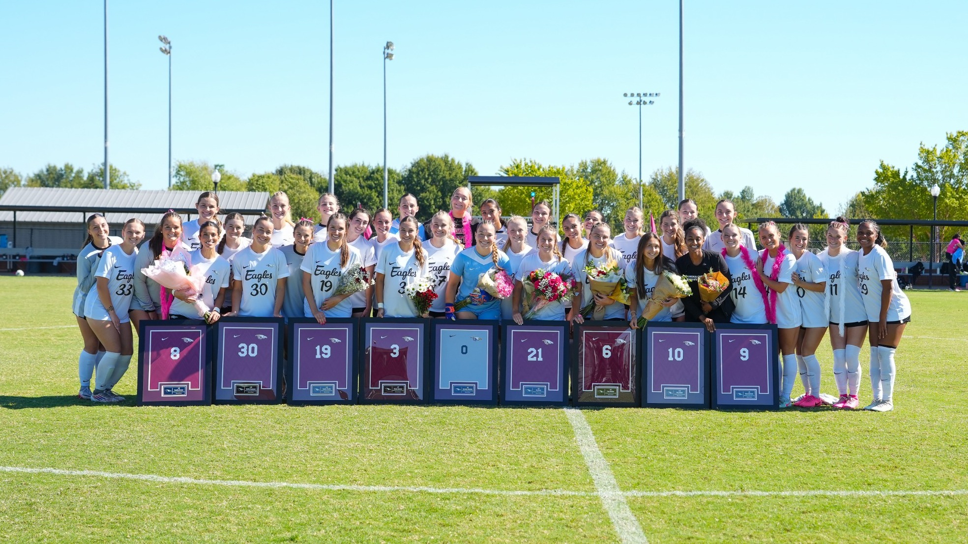 The Oklahoma Christian women's soccer team poses together on the field during a sunny Senior Day celebration. The nine senior players stand near the front holding flower bouquets, positioned behind framed maroon and light blue jerseys displaying their numbers (8, 30, 19, 3, 0, 21, 6, 10, and 9). Teammates stand alongside them on the grass, with light poles and trees lining the background.
