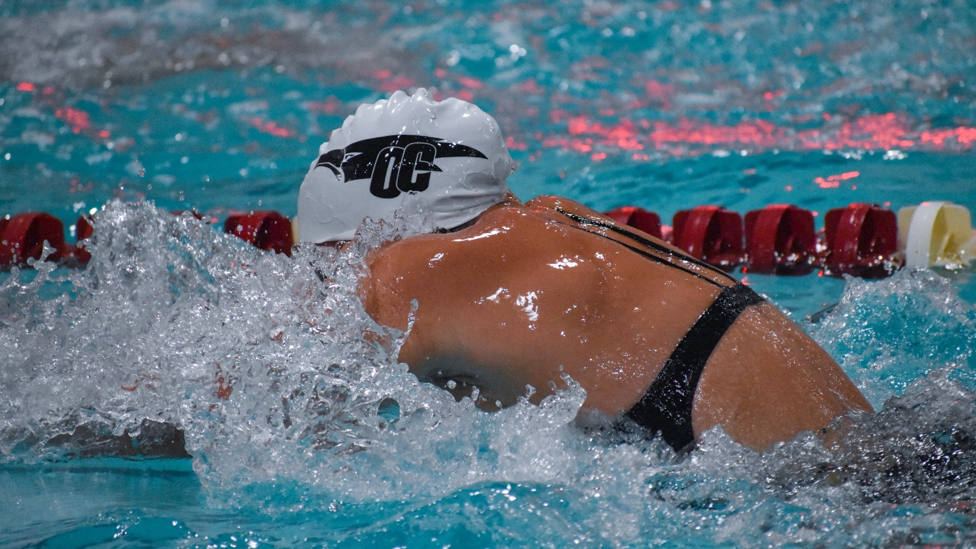 Oklahoma Christian Women's Swimming Competes Swimming in a black suit with white cap featuring a black OC logo