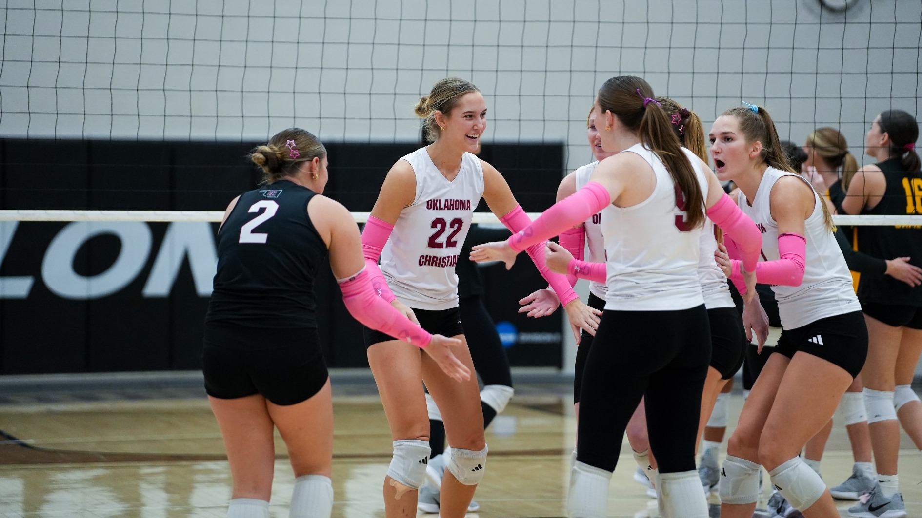 Oklahoma Christian volleyball players in white jerseys and pink arm sleeves huddle and celebrate at the net. Chloe Jones (22) smiles at the center of the group while libero Macey Nehring (2), in a black jersey, reaches toward her. Cameron players in black and gold stand in the background.