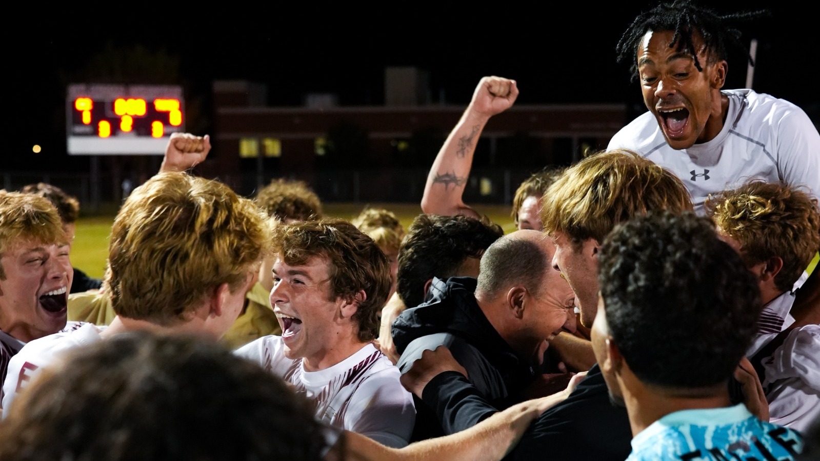 Oklahoma Christian men’s soccer players and staff celebrate on the field at night after the win, shouting and pumping their fists as they crowd around a man in a black jacket. A goalkeeper in a bright blue Eagles jersey reaches in, and the lit scoreboard glows in the background.