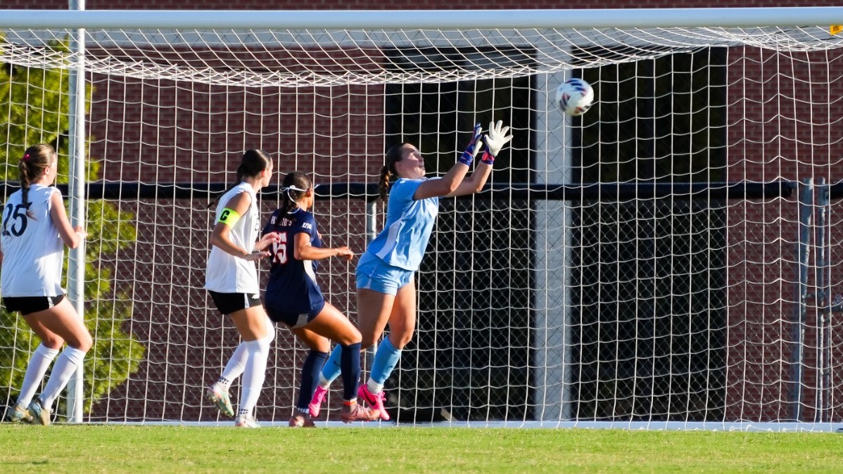 On a sunny afternoon, an Oklahoma Christian goalkeeper in a light blue kit stretches both gloved hands up toward a ball dropping into the goalmouth. Three OC teammates in white jerseys, including one wearing a captain’s armband, stand around her.