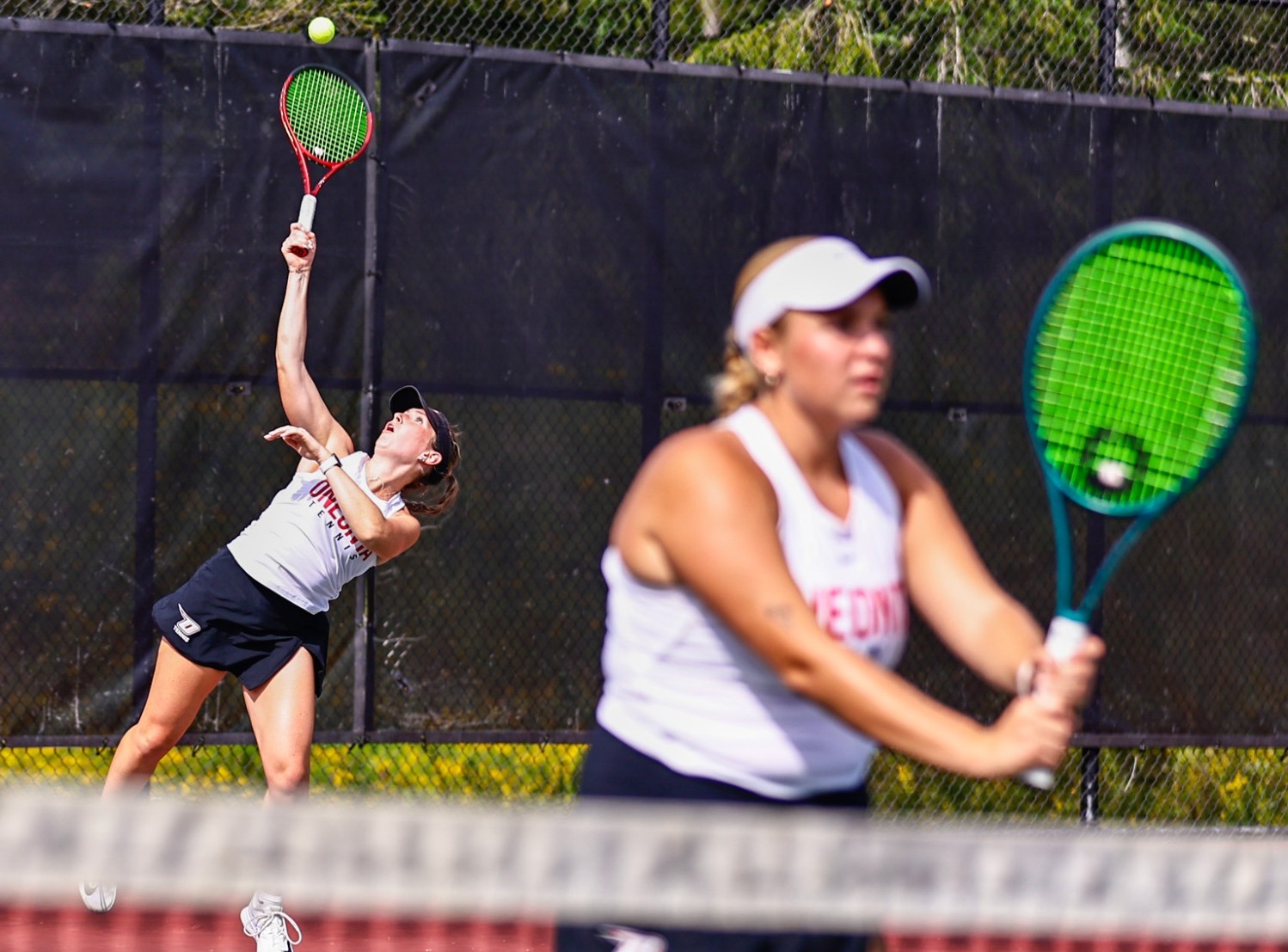 Annika Cripe serving in doubles play with Haley Peck in the front against Hartwick