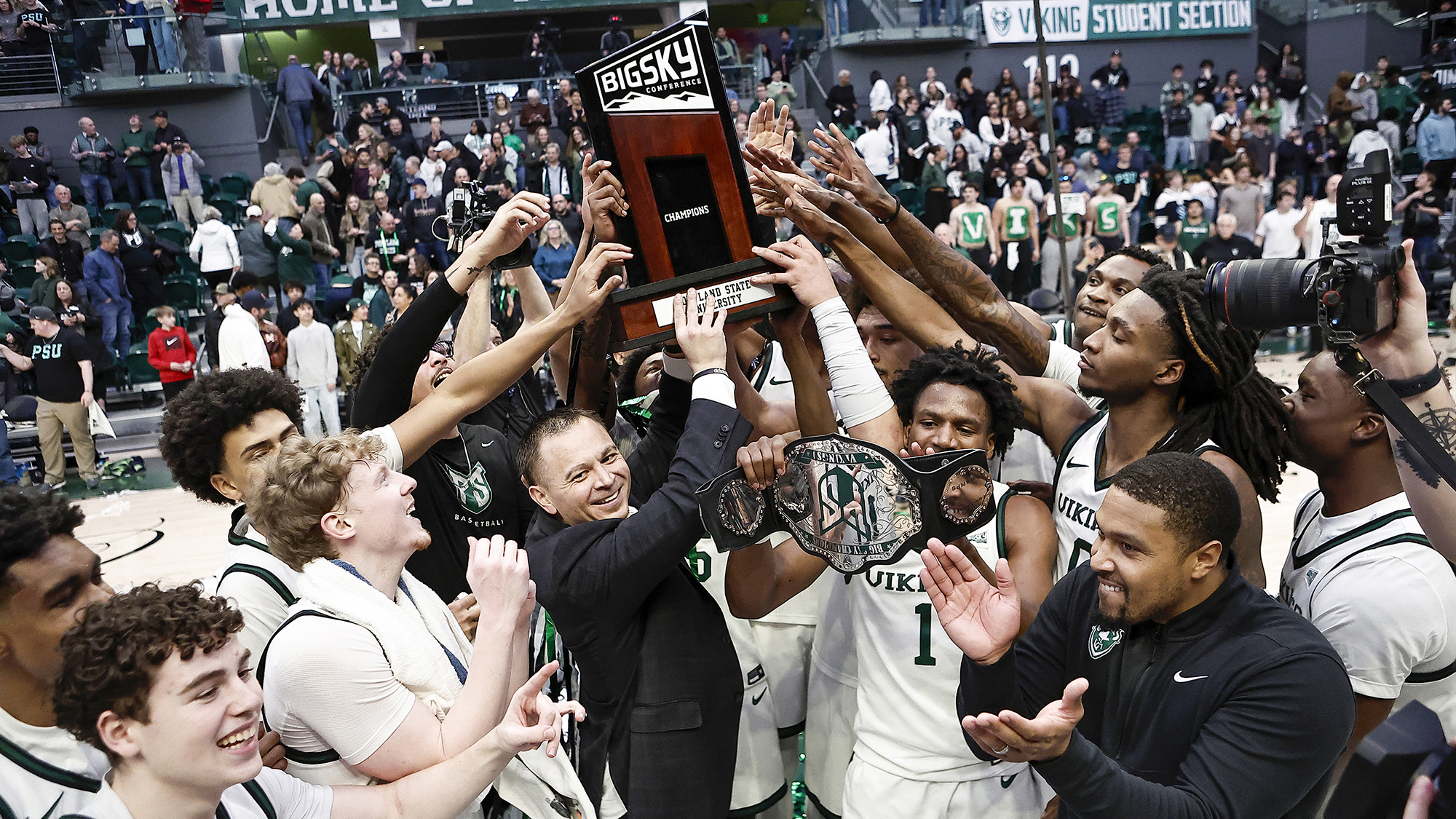 Portland State Head Coach Jase Coburn holds aloft the 2025-26 Big Sky Championship trophy while surrounded by his team.