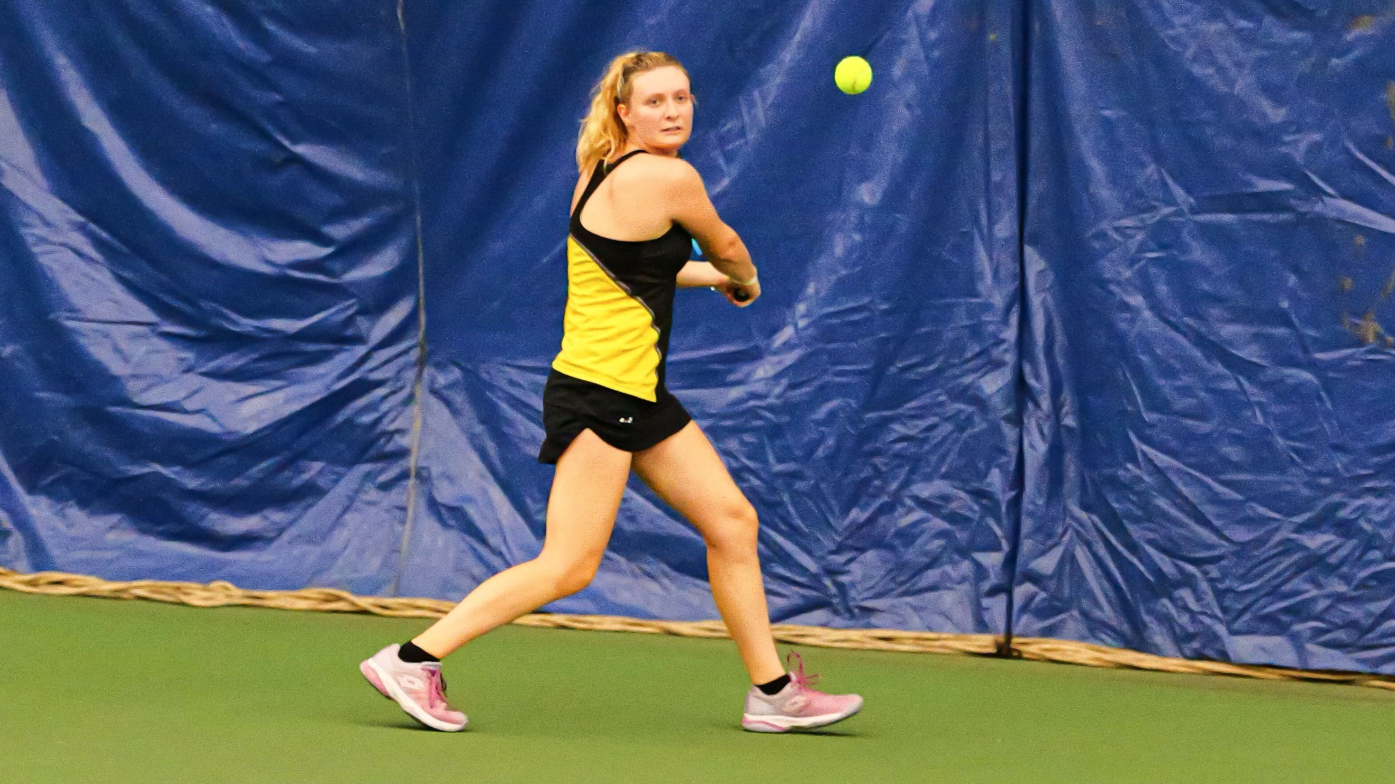 Beatrice Crespo in a black and yellow tank top and shorts on a green court, tracking a yellow tennis ball mid-air to hit a backhand against a blue backdrop.   
