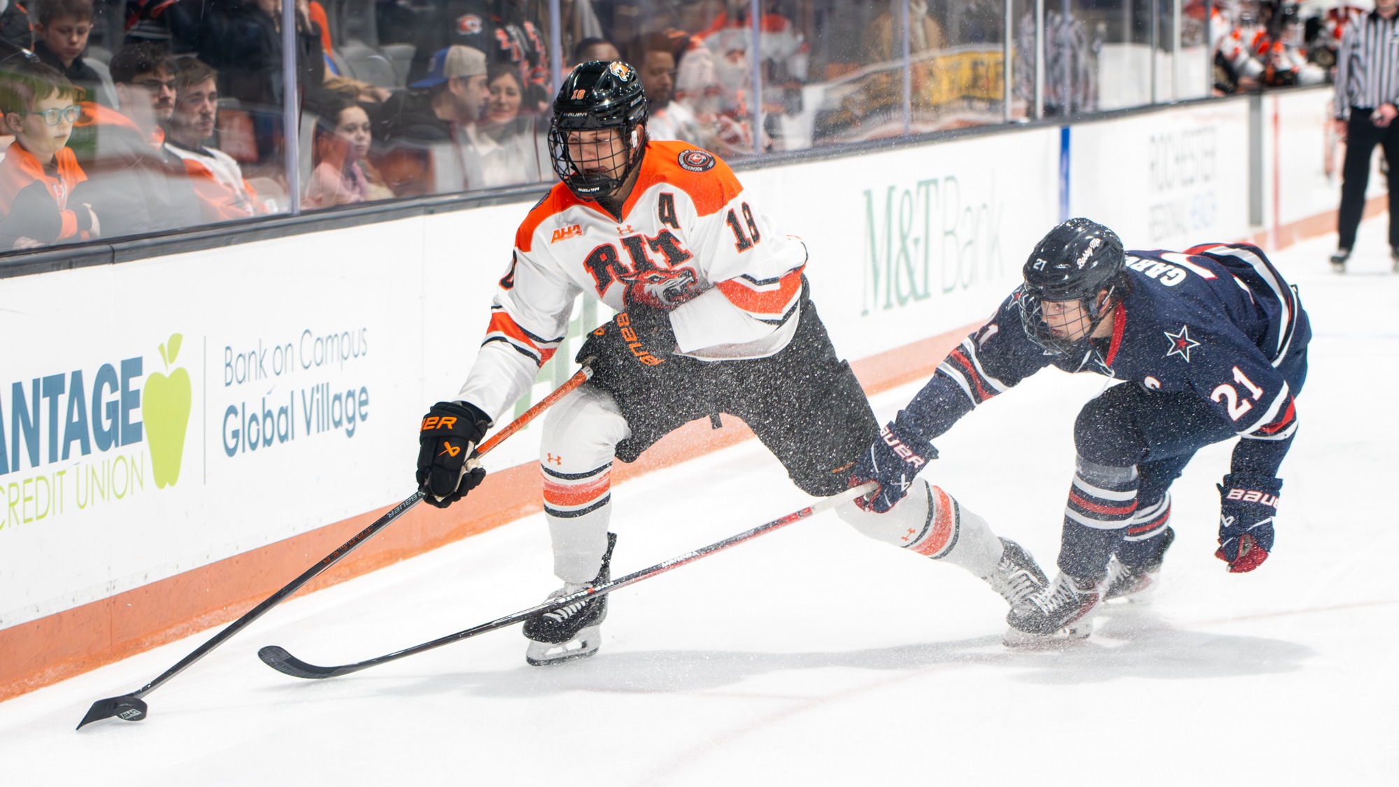 a men's hockey player skates with the puck along the boards ahead of a defenseman