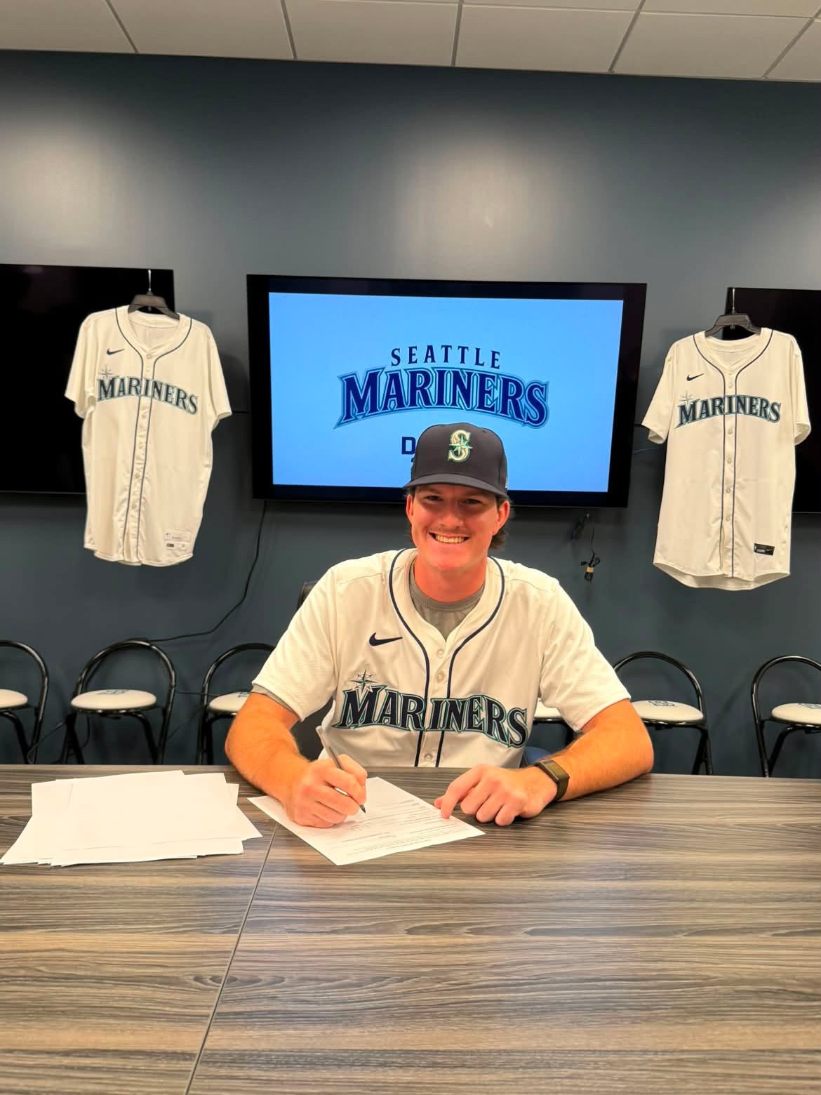 Isaac Thornton wearing a Seattle Mariners baseball jersey and cap smiling at the camera while sitting at a desk and holding a pen to sign official contract papers. In the background, a TV screen displays 