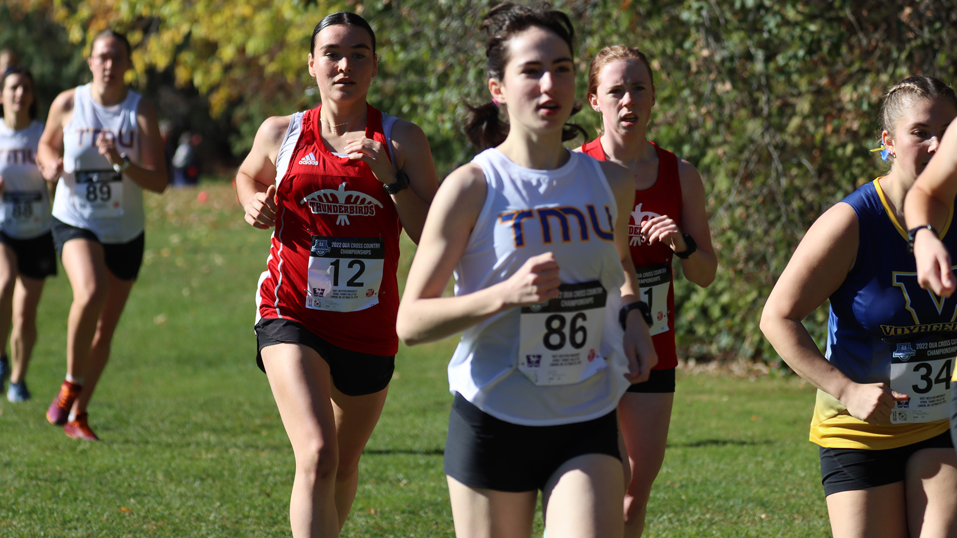 A TMU runner races in a small pack with four other runners, while another TMU runner follows in the background.