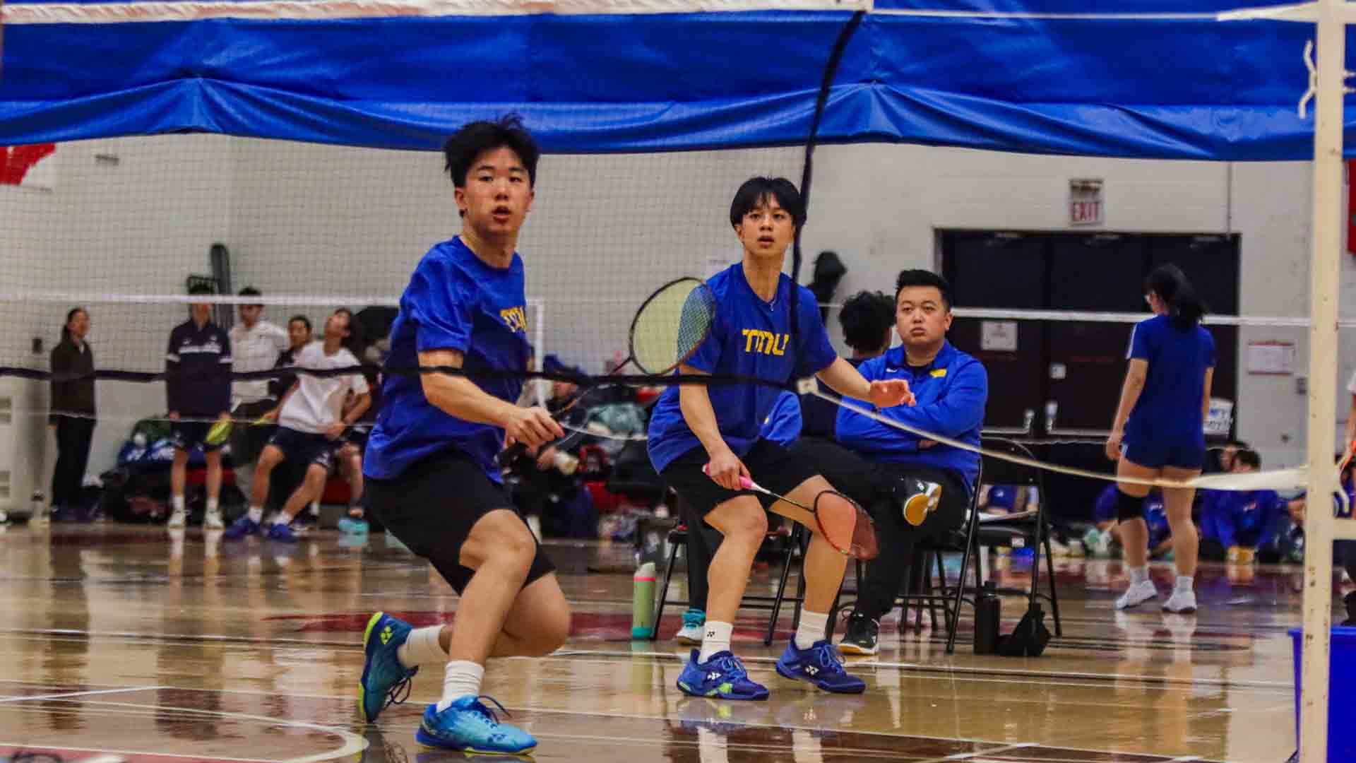 Looking through the net at two TMU Bold men's badminton players in action
