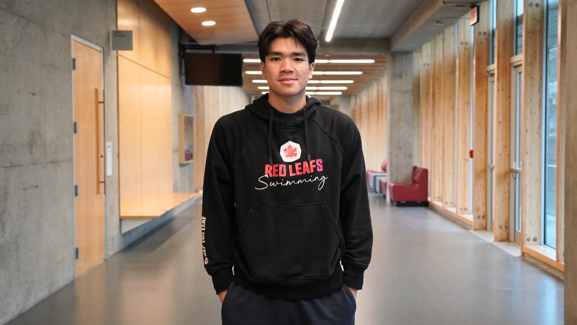 Marcus Mak in an SFU Swimming hoodie, standing in Blusson Hall on campus.