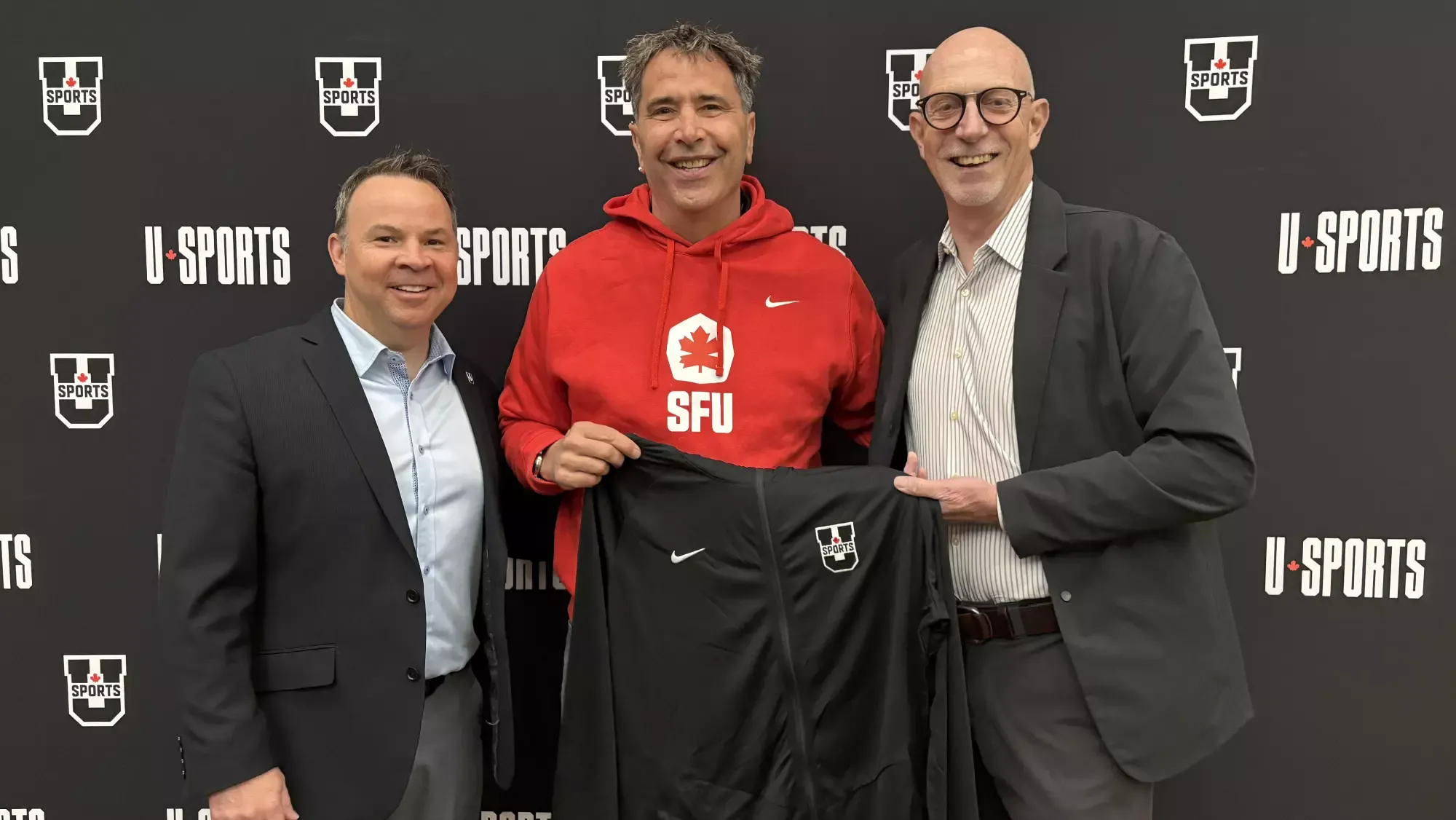 SFU Executive Director Luc Simard smiles alongside U SPORTS members while wearing an SFU sweatshirt and holding up a U Sports jacket