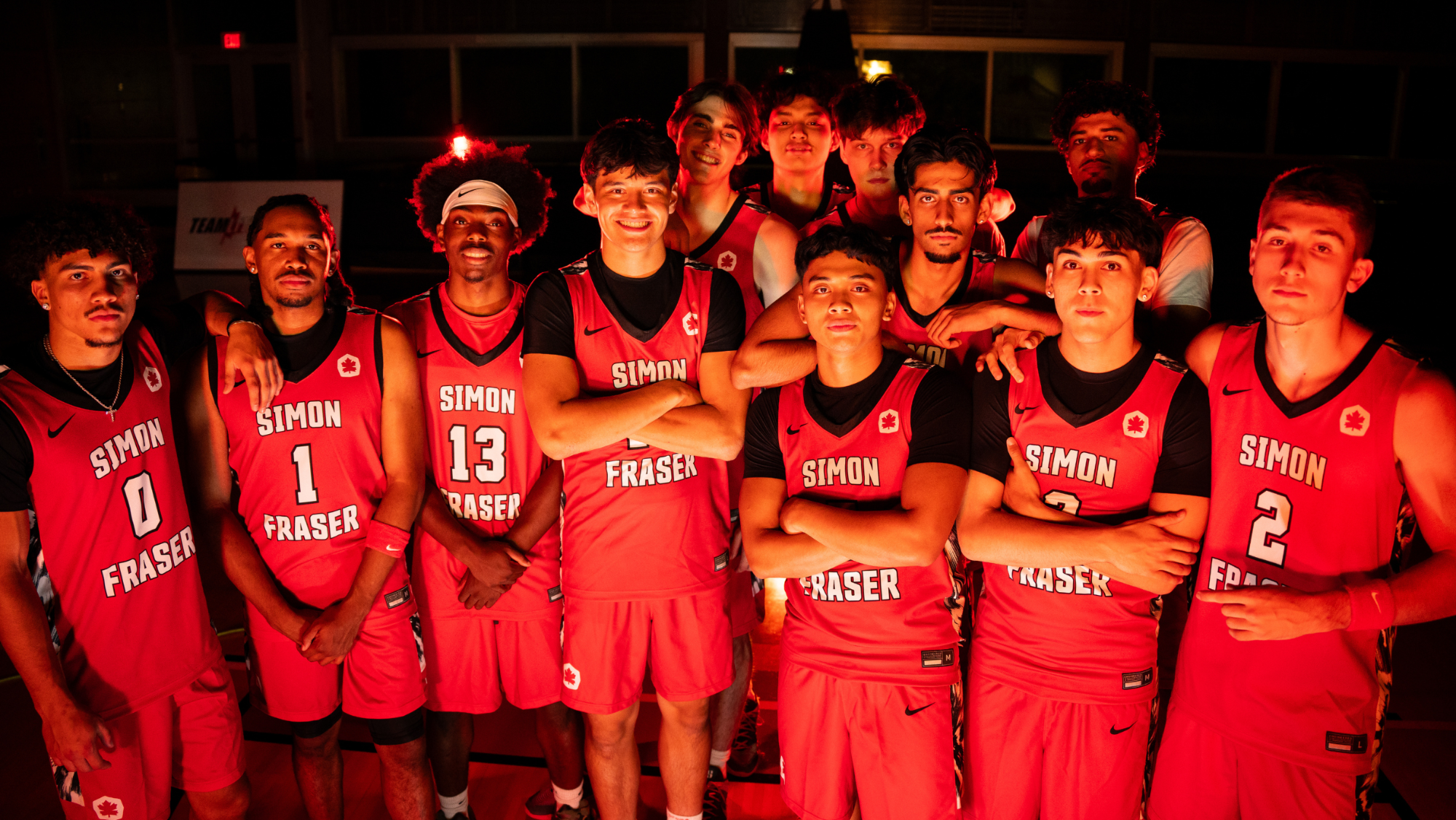 The SFU men's basketball team poses together on media day. In red uniforms. Lit from the front with a dark background. 