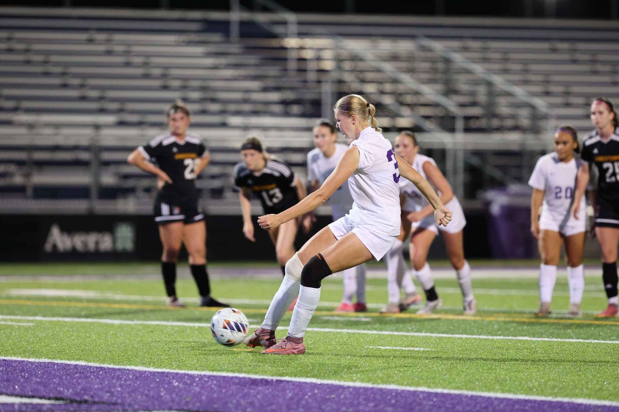 A women’s soccer player in white strikes the ball on a brightly lit field as opponents and teammates watch.