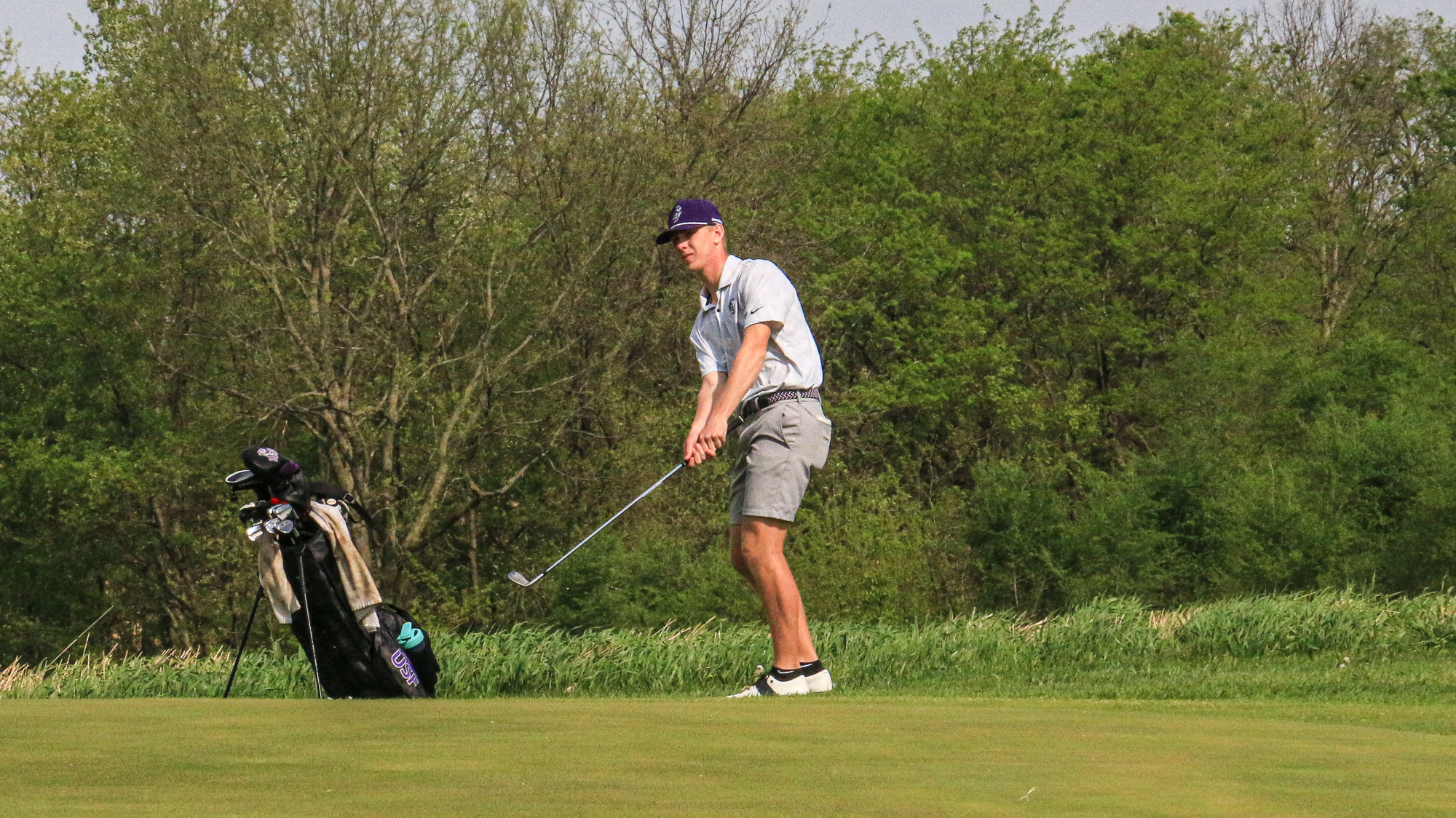 Charlie Hanson chips toward the green during tournament play, with his golf bag standing nearby and trees lining the course in the background.