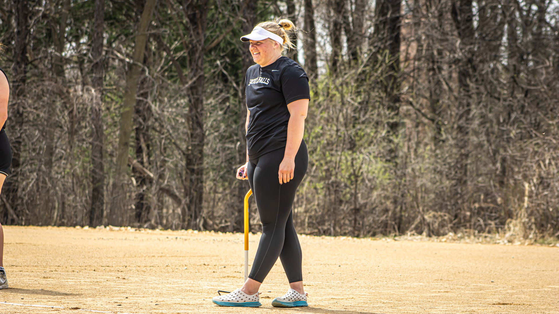 Delanie Van Driel walking across a throwing area during a track and field meet, wearing a black Sioux Falls shirt, black leggings, white visor, and light-colored Crocs, with a wooded background behind her.
