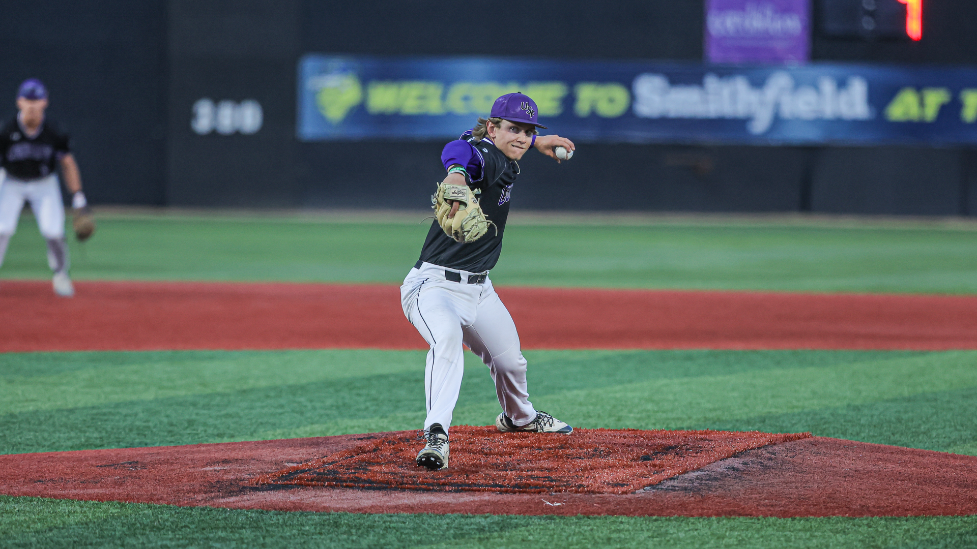 University of Sioux Falls pitcher Zach Peta delivers a pitch from the mound at Sioux Falls Stadium, with a fielder ready behind him during game action.