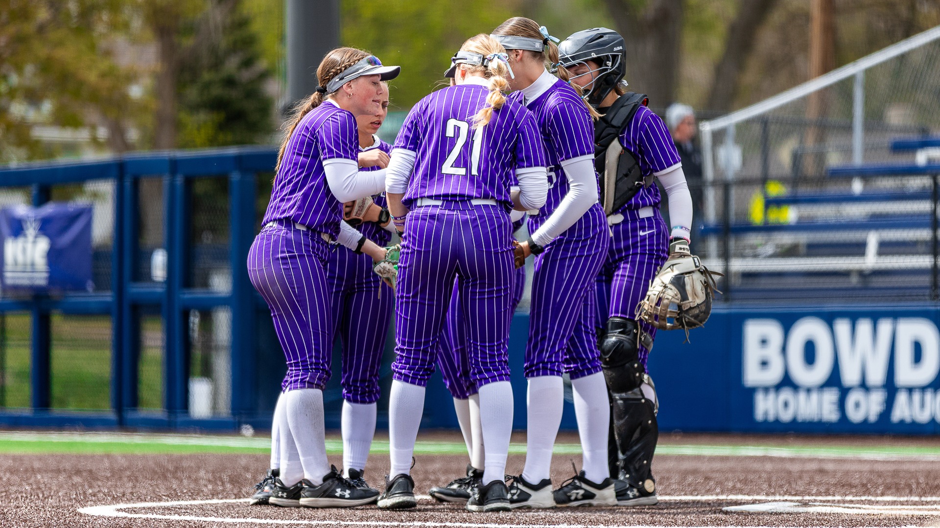 USF softball players gather in a tight circle on the pitcher’s mound during the NSIC Tournament at Bowden Field, wearing purple pinstripe uniforms as they meet to regroup mid-inning.