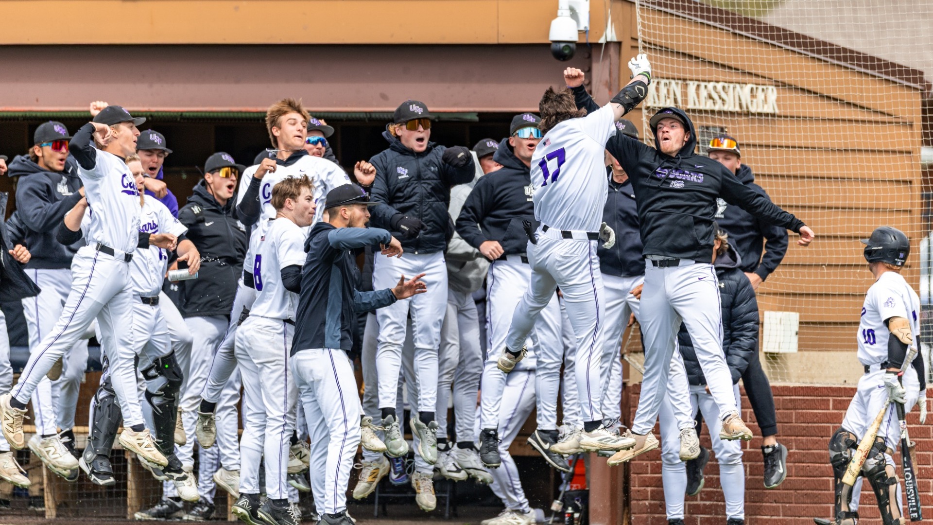 Mitch Iliff leaps into a mob of celebrating Sioux Falls teammates near the dugout after hitting a home run, as players jump and cheer in excitement along the fence line.