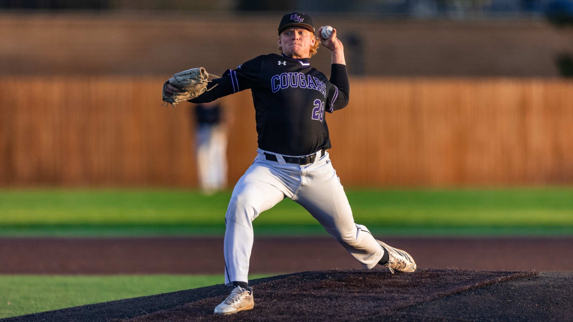 Owen Weadge delivers a pitch from the mound during a game at Ronken Field, wearing a black Sioux Falls Cougars jersey, captured mid-throw with strong form and focus.