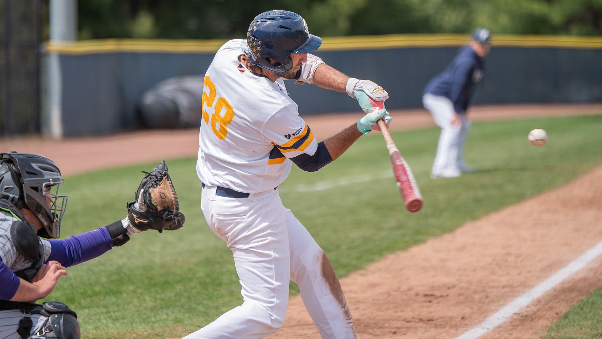 Jake Pisano swinging at a pitch, with the catcher in the foreground and head coach Chris Shank in the background