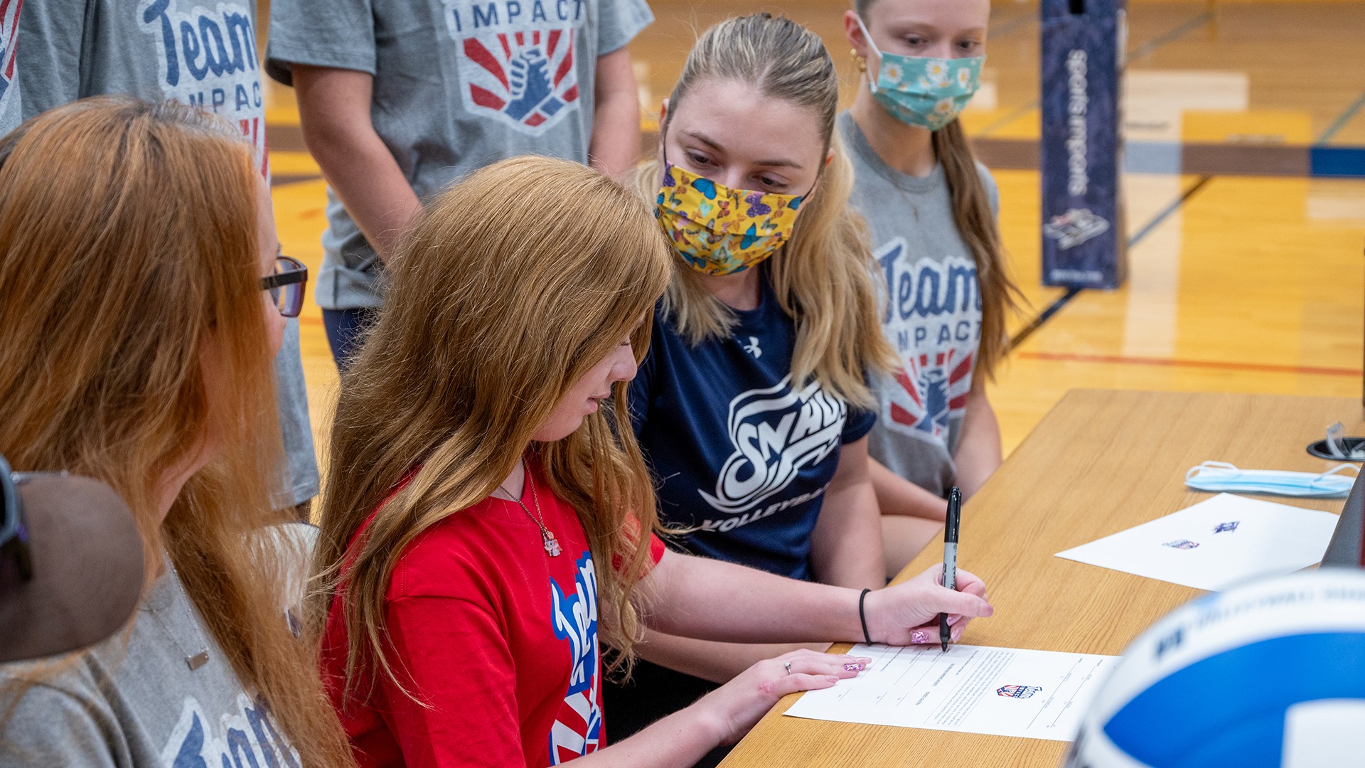 Madison McCann signing her Letter of Intent to join the SNHU volleyball team after matching through Team IMPACT, while head coach Lauren Havill and teammates look on