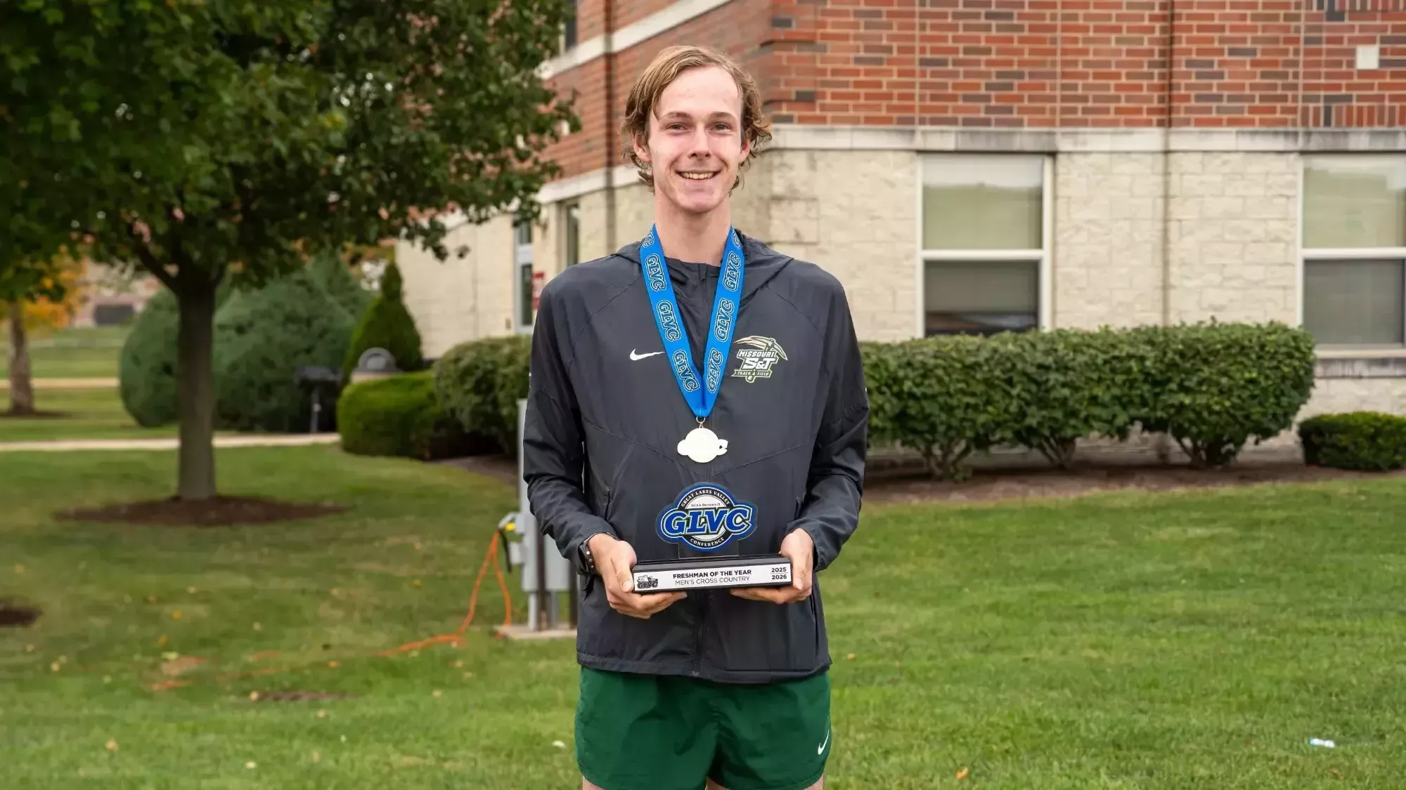 Maddox Riley photographed smiling and holding a GLVC trophy for winning Freshman of the Year. He is wearing a gray warmup jacket and has a medal around his neck.