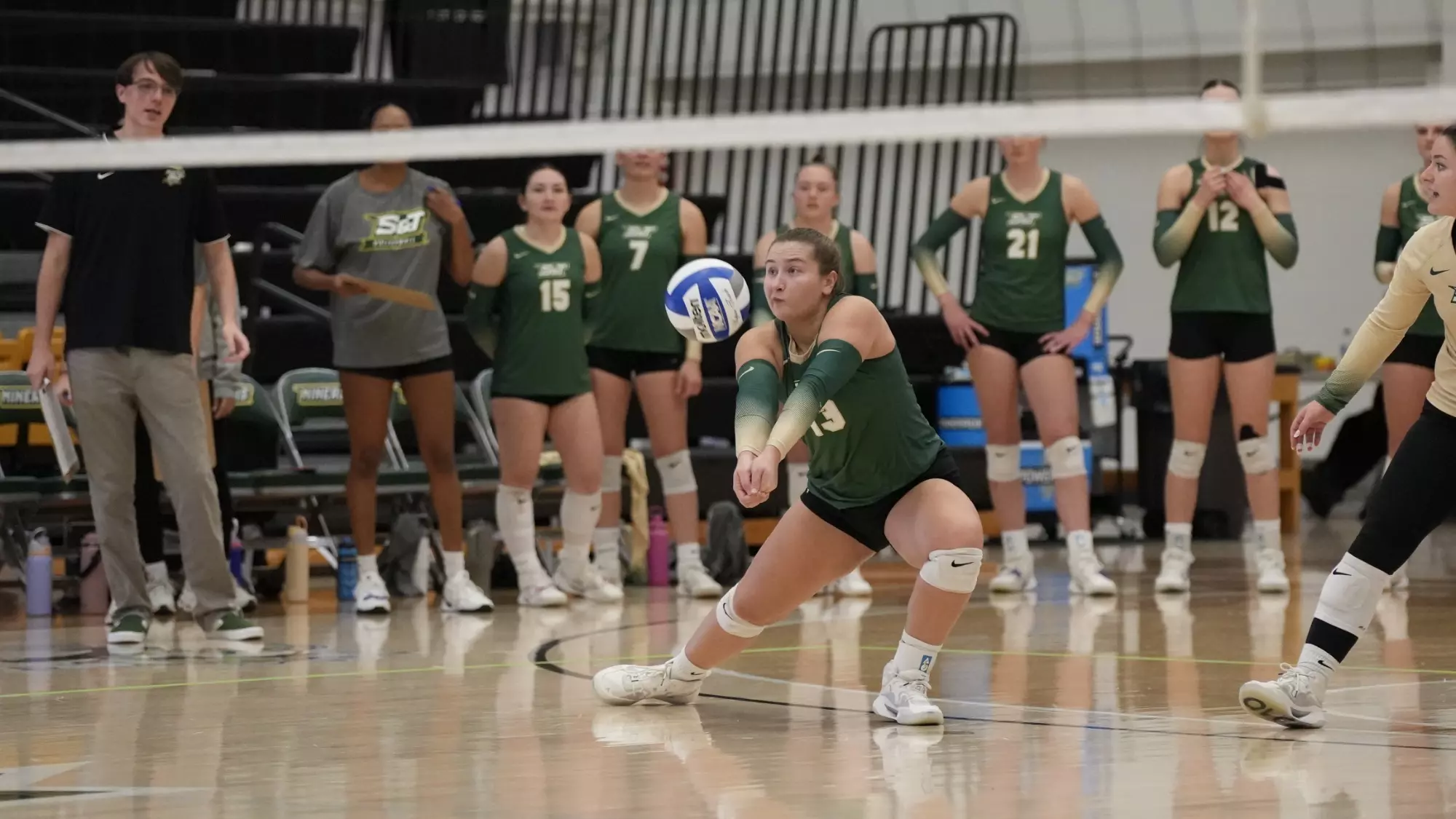 A photo of Lexi Kosmopolis wearing a green jersey (13) at mid court making a play at a volleyball that was hit in her direction. S&T volleyball players can be seen in the background standing in front of the bench.