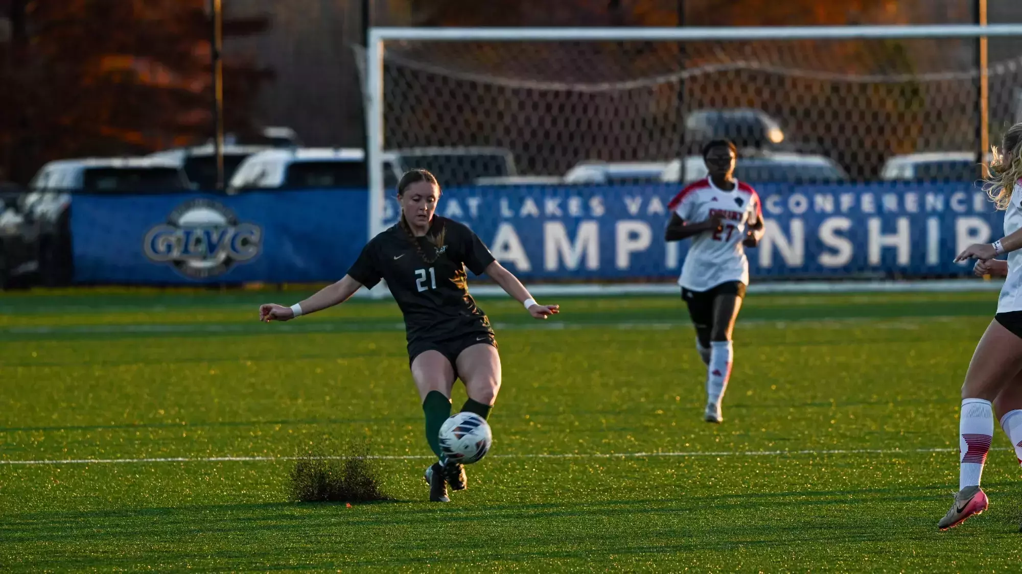 Women's soccer player Sarah Henze wearing a dark green jersey (21) dribbling a ball upfield. A Drury defender can be seen in the background along with the GLVC Championship banner behind the Drury goal.