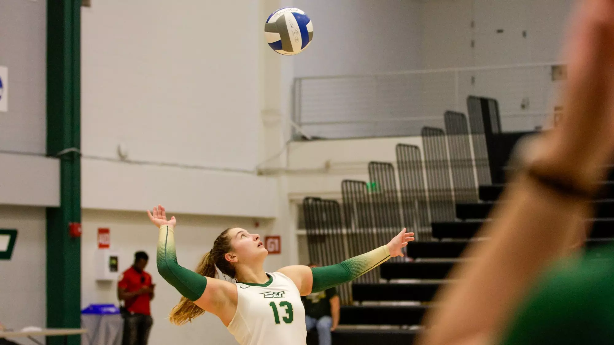 A photo of Lexi Kosmopolis wearing a white jersey (13) in the middle of her serve, arm raised ready to hit the volleyball that is suspended above her head.