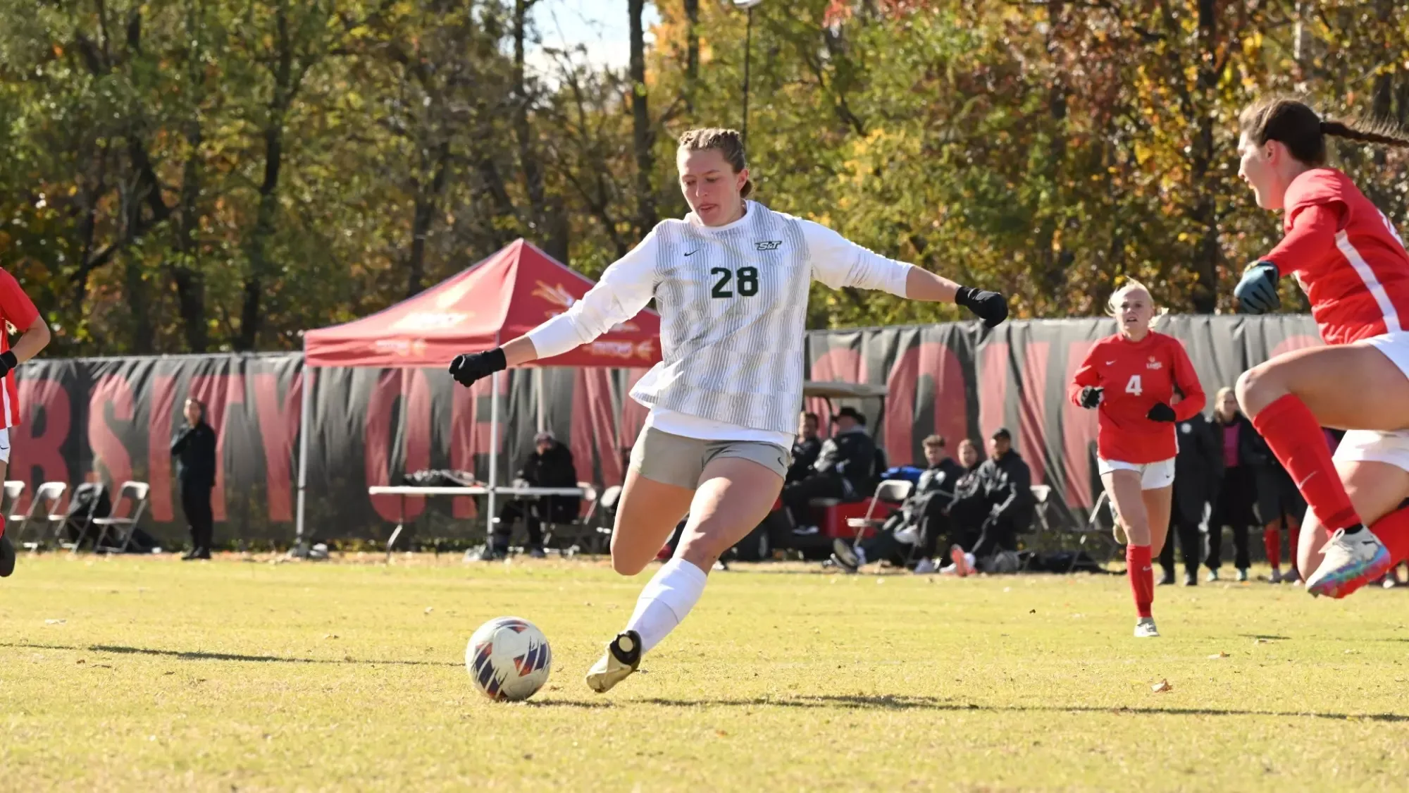 Adriana Benassi of Missouri S&T's women's soccer photographed mid-kick during a match against UMSL as she attempts to score a goal. She is wearing a white uniform top with matching light gray shorts.