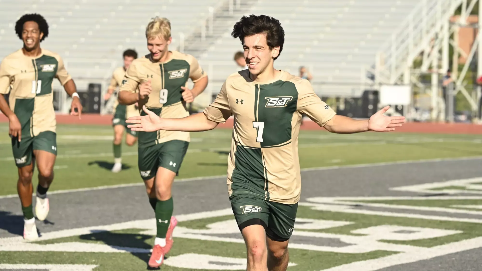 Missouri S&T's Joao Vieira photographed celebrating after a goal during a match at Allgood-Bailey Stadium. He holds his hands out in celebration and wears a green and gold checkered uniform with green shorts