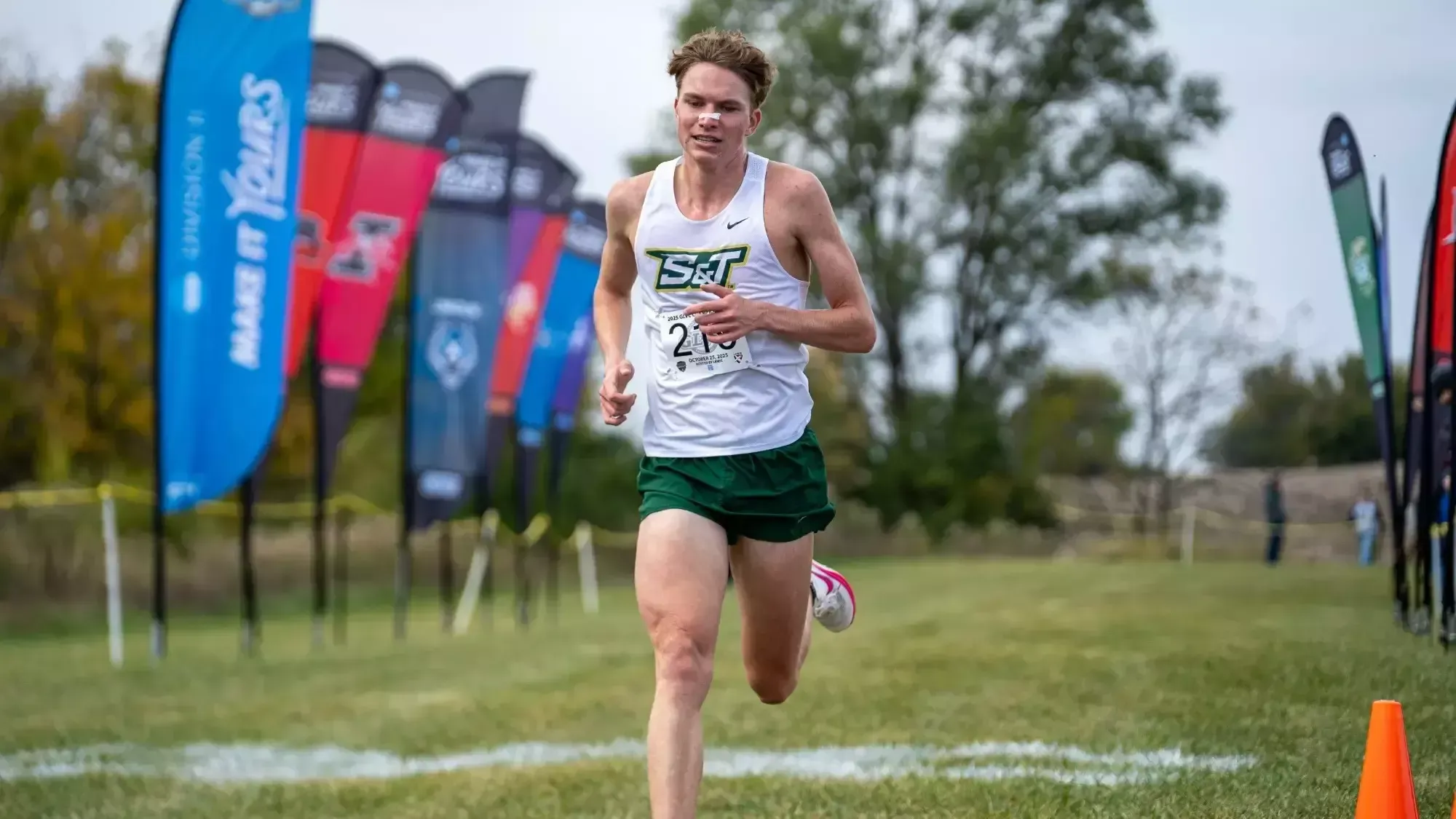 Henry Born photographed running in stride during the GLVC cross country championships. He is wearing a uniform with a white top and green shorts.