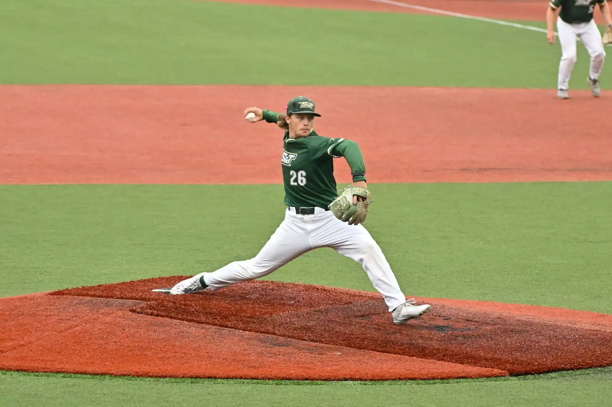 Jake Schisler of missouri S&T baseball photographed throwing a pitch during the GLVC tournament. He is wearing a green uniform top and white pants as he steps with his arm up to deliver a pitch