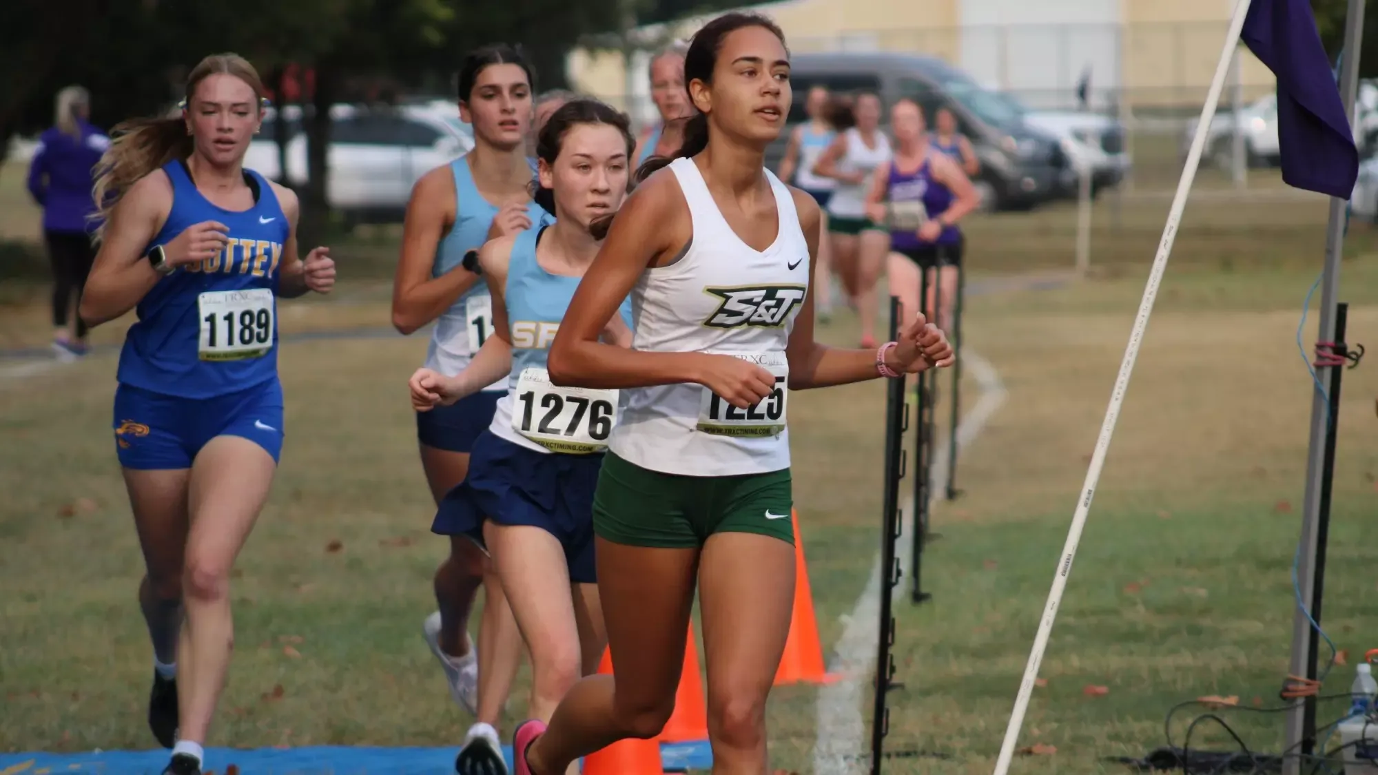 Grace Johnson of S&T cross country photographed running in stride during a meet at the Bearcat Invite. She is wearing a white top with green shorts