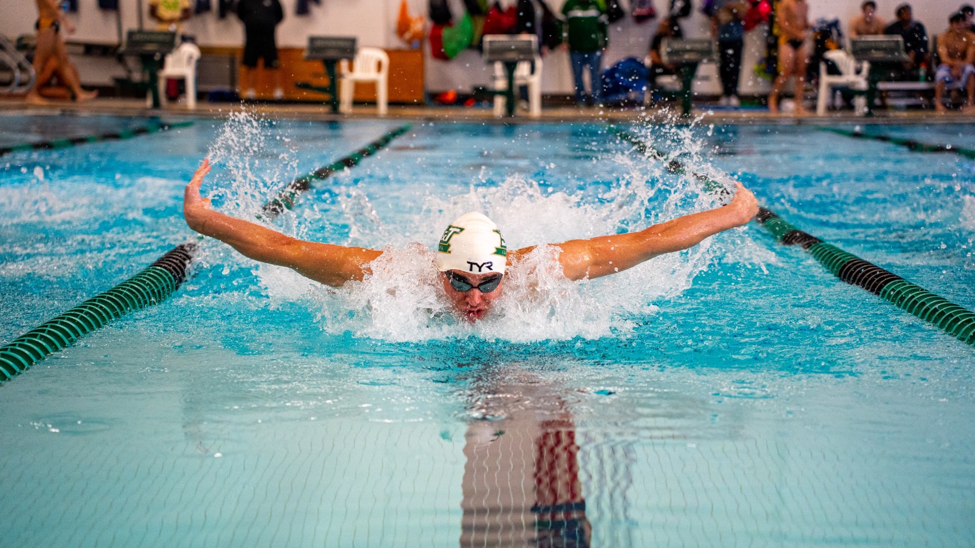 A competitive swimmer performing the butterfly stroke in an indoor pool. The swimmer has both arms extended wide out of the water and splashing spray around them. They are wearing a white swim cap with a green logo, black goggles, and are facing forward. The background shows pool lanes, starting blocks, and spectators on the deck.