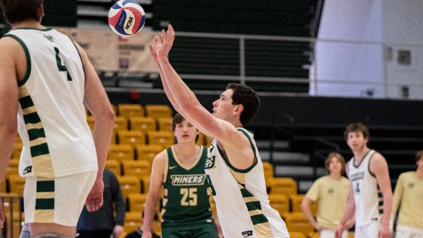 Zachary Carff of Missouri S&T Men's Volleyball wearing green jersey and digs the volleyball with both hands during a men’s volleyball match, as teammates prepare to attack and opponents stand ready across the net, with the court and bench visible in the background.