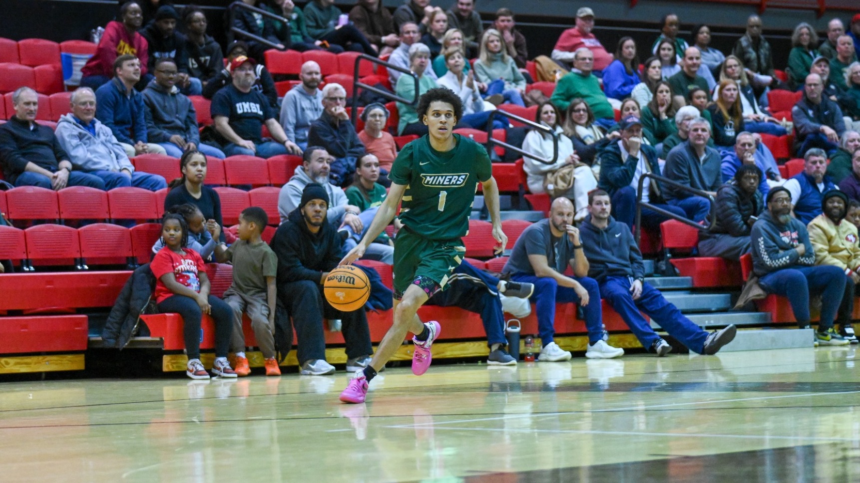 Missouri S&T men's basketball player Blaise Beauchamp wearing a green jersey and dribbling a basketball with the stands and crowd in the background at a basketball game at Missouri-St. Louis
