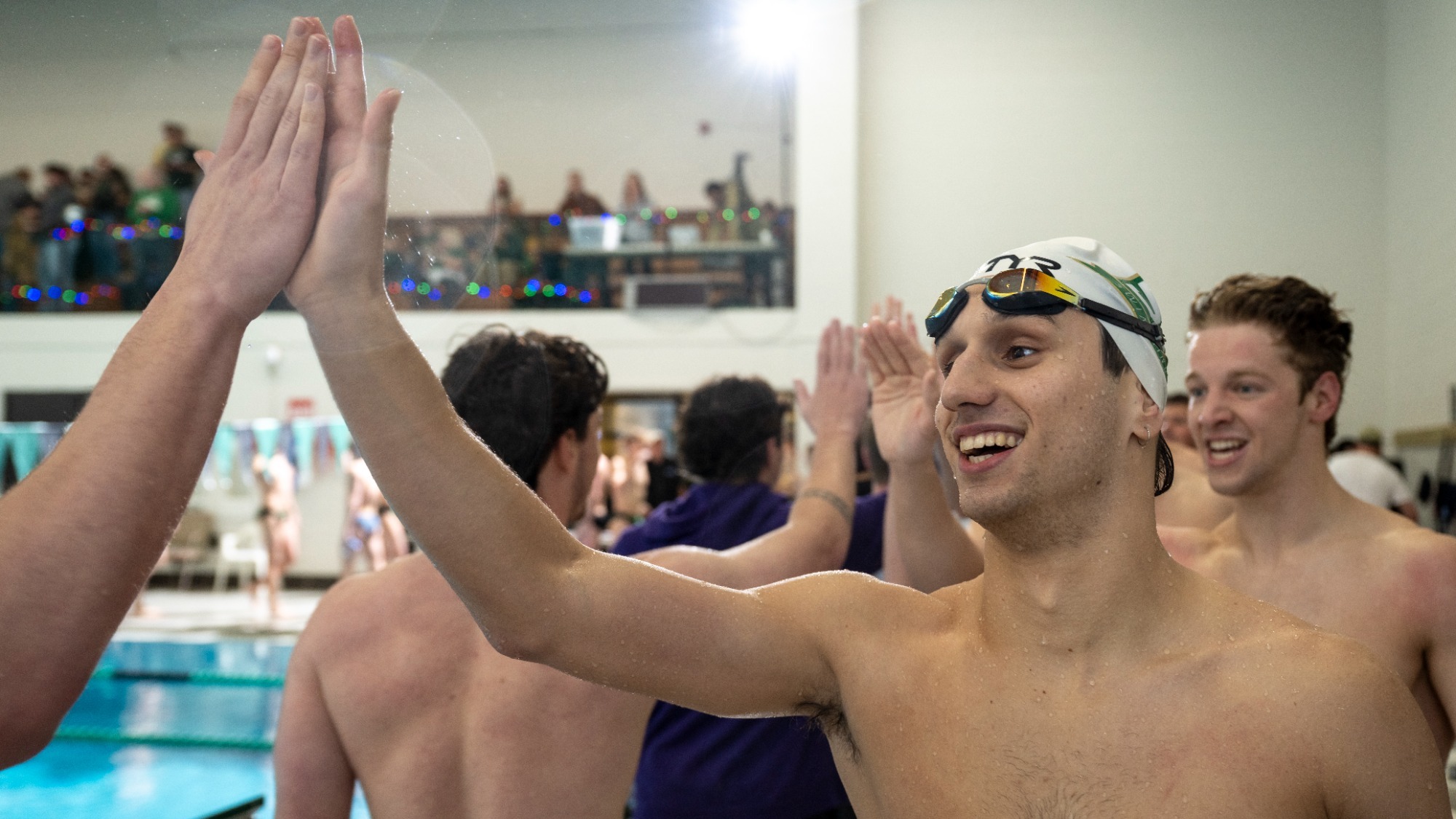 Missouri S&T swimmer Giorgio Panci smiles and gives a high-five to an opposing teammate at the edge of a competition pool. Panci wears a white swim cap and goggles, with other cheering teammates visible in the background.