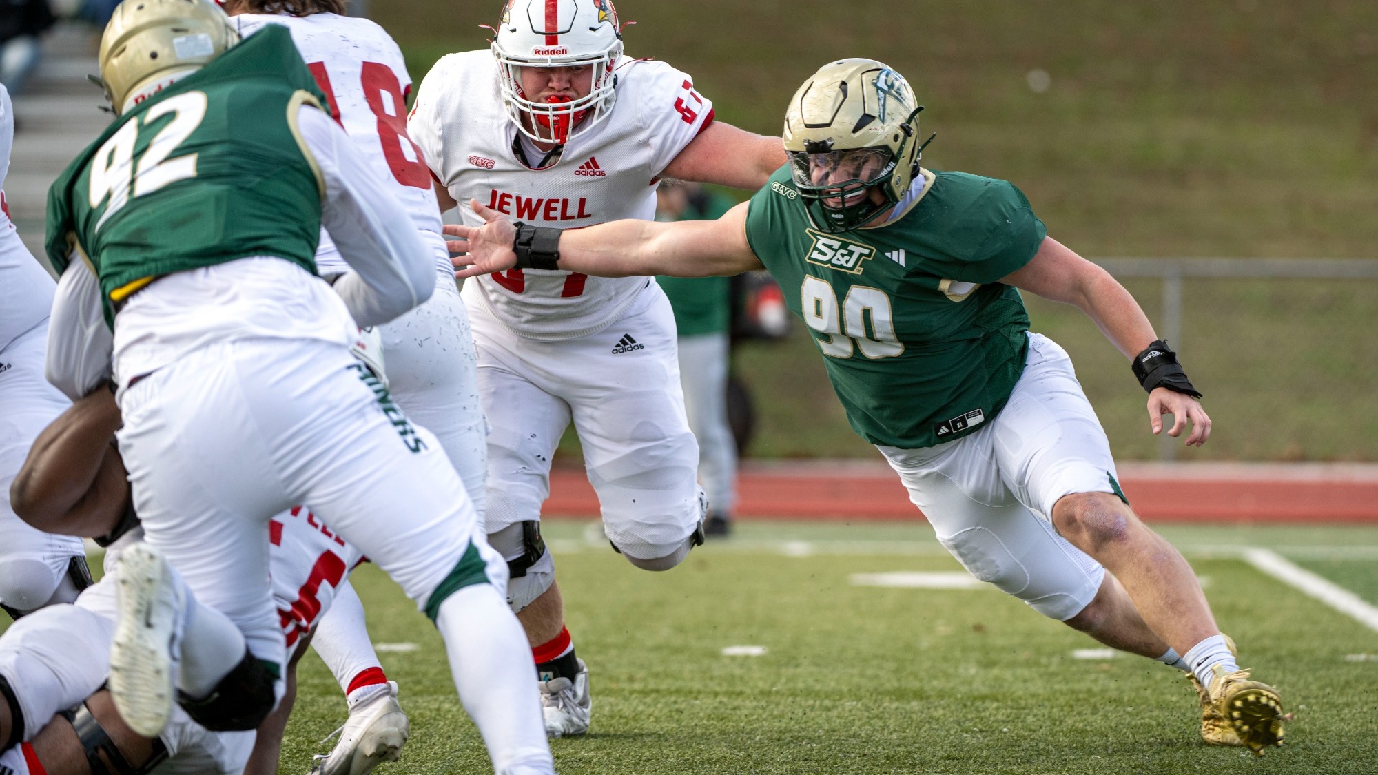Missouri S&T football athlete Bentley Hart wearing a green and white jersey with the number 90 on it, running towards an offensive player on the William Jewell Cardinals with his arm outstretched to make a tackle